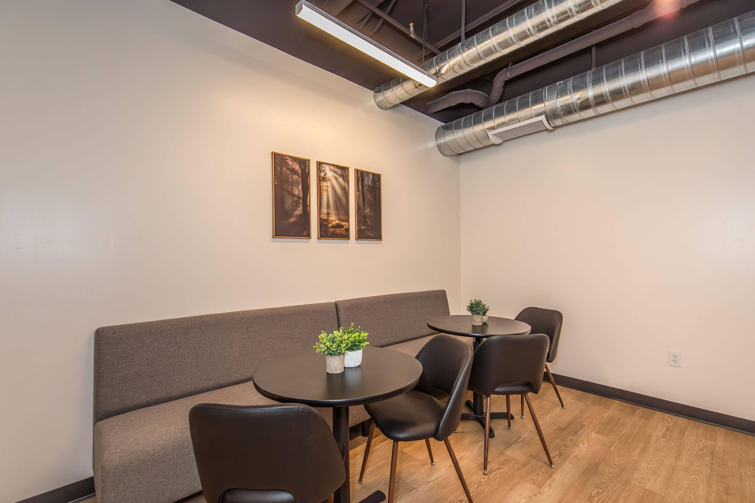 A modern, minimalist coffee shop interior featuring two round black tables with four black chairs. A gray couch lines the wall adorned with two framed nature photographs. A small green plant decorates each table, and the ceiling has exposed ducts, adding a contemporary touch to the space.