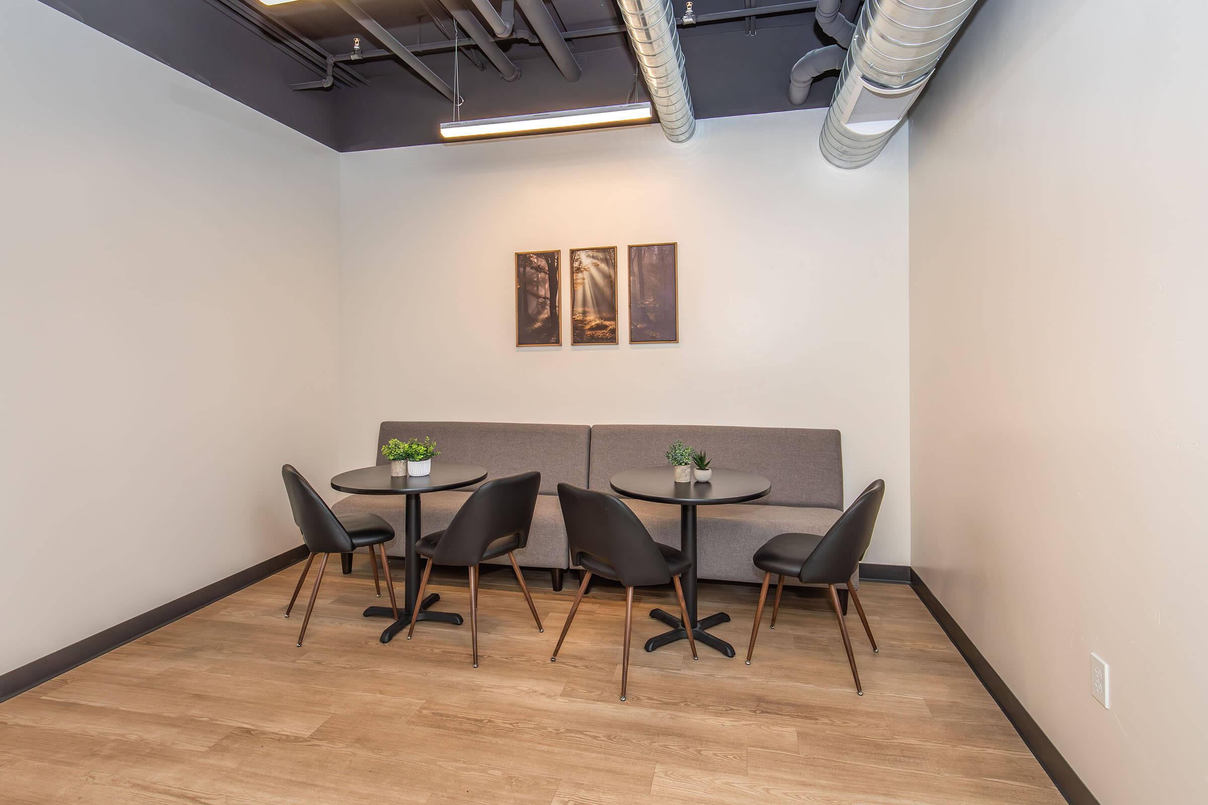 Cozy corner of a modern space featuring a gray sofa against the wall, two framed pictures above it, and two round black tables surrounded by black chairs. Small potted plants adorn each table, and the flooring is light wood, giving a warm and inviting atmosphere.