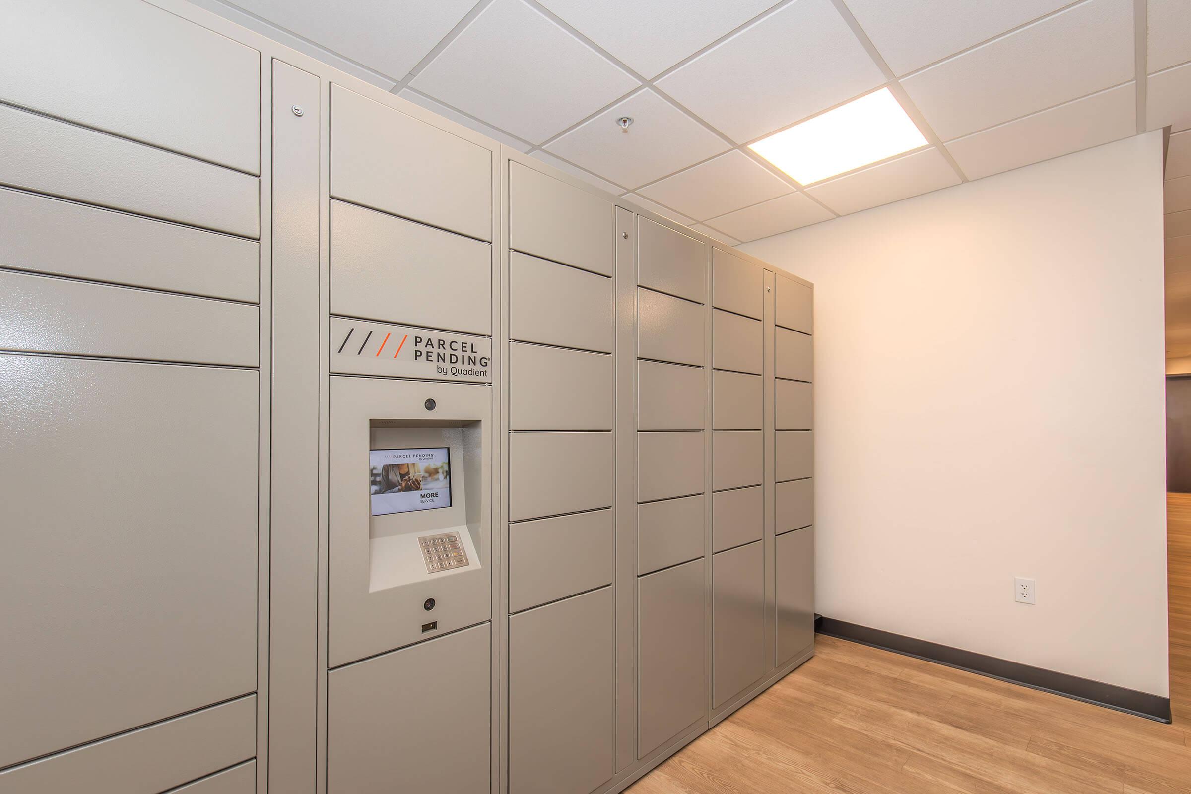 Indoor parcel locker system against a wall with multiple compartments. A touchscreen interface is visible for package retrieval, and the area is well-lit with a modern aesthetic. The flooring is wood-like, complementing the neutral color scheme of the space.