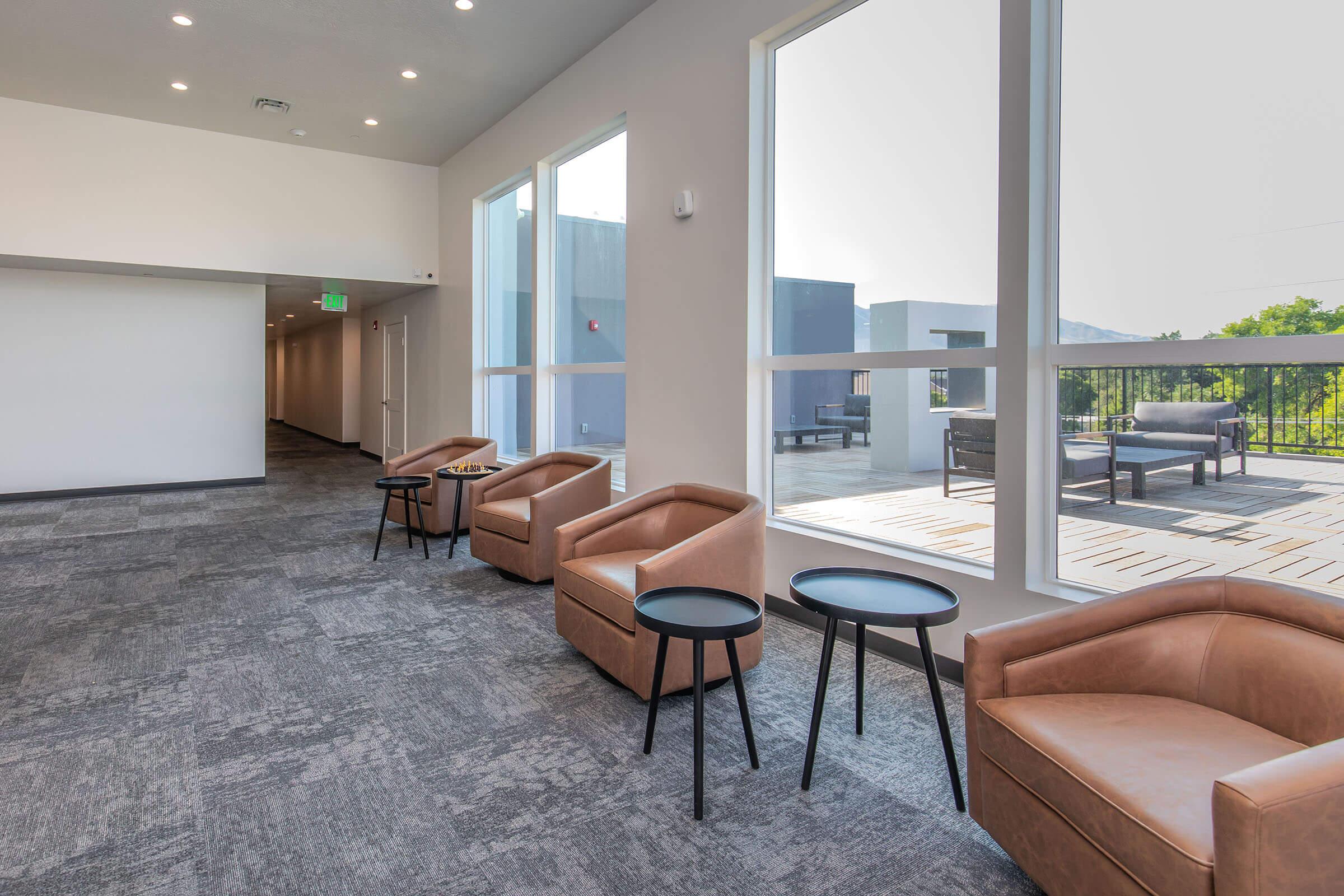 A modern, well-lit lounge area with large windows letting in natural light. There are several brown upholstered chairs paired with small black tables. The space features a contemporary design and leads to an outdoor area visible through the windows.