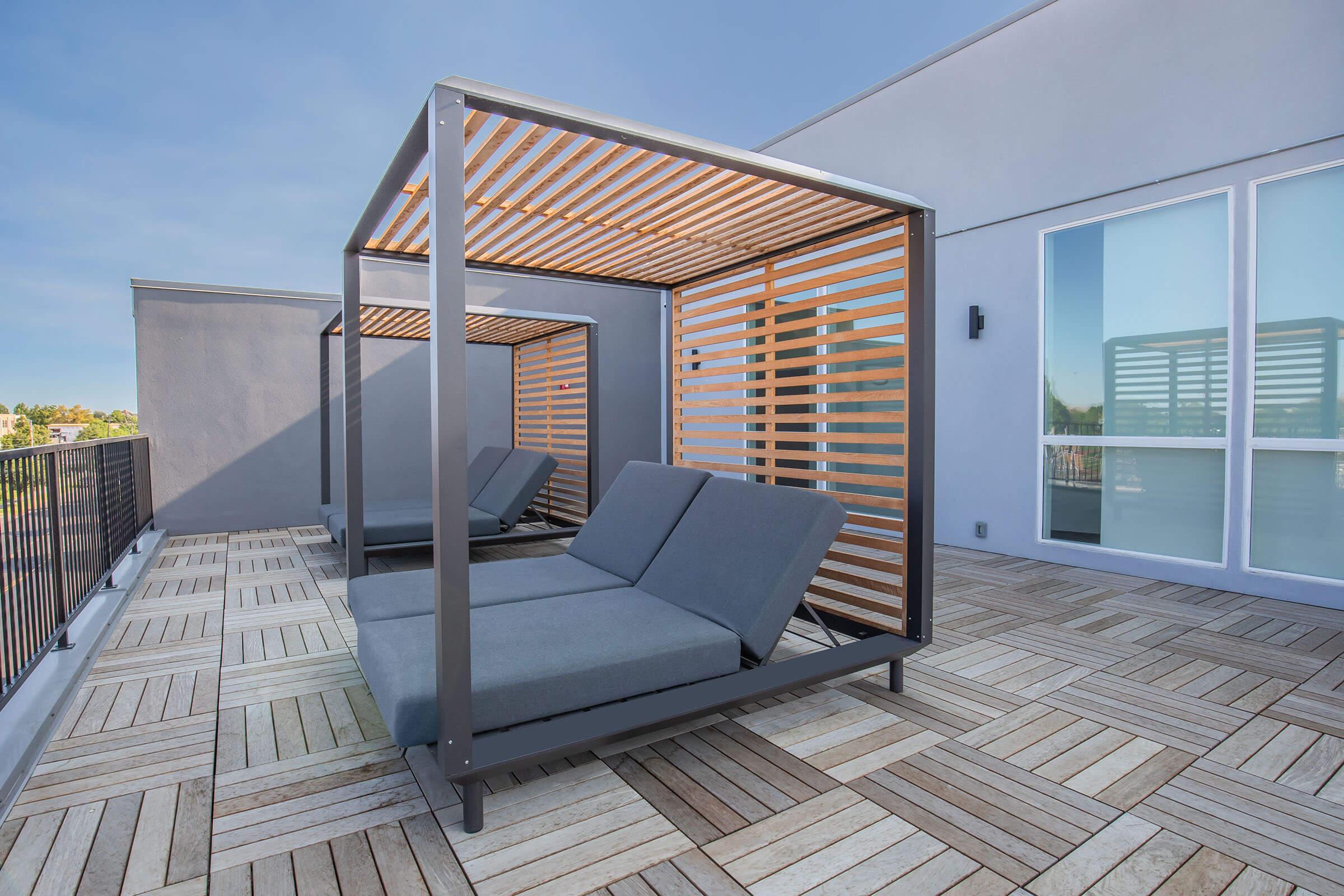 Two modern outdoor lounge spaces featuring metal frames with wooden slats, situated on a rooftop terrace. The area has a spacious tiled floor and a clear blue sky in the background, creating a relaxing atmosphere for lounging or enjoying the view.