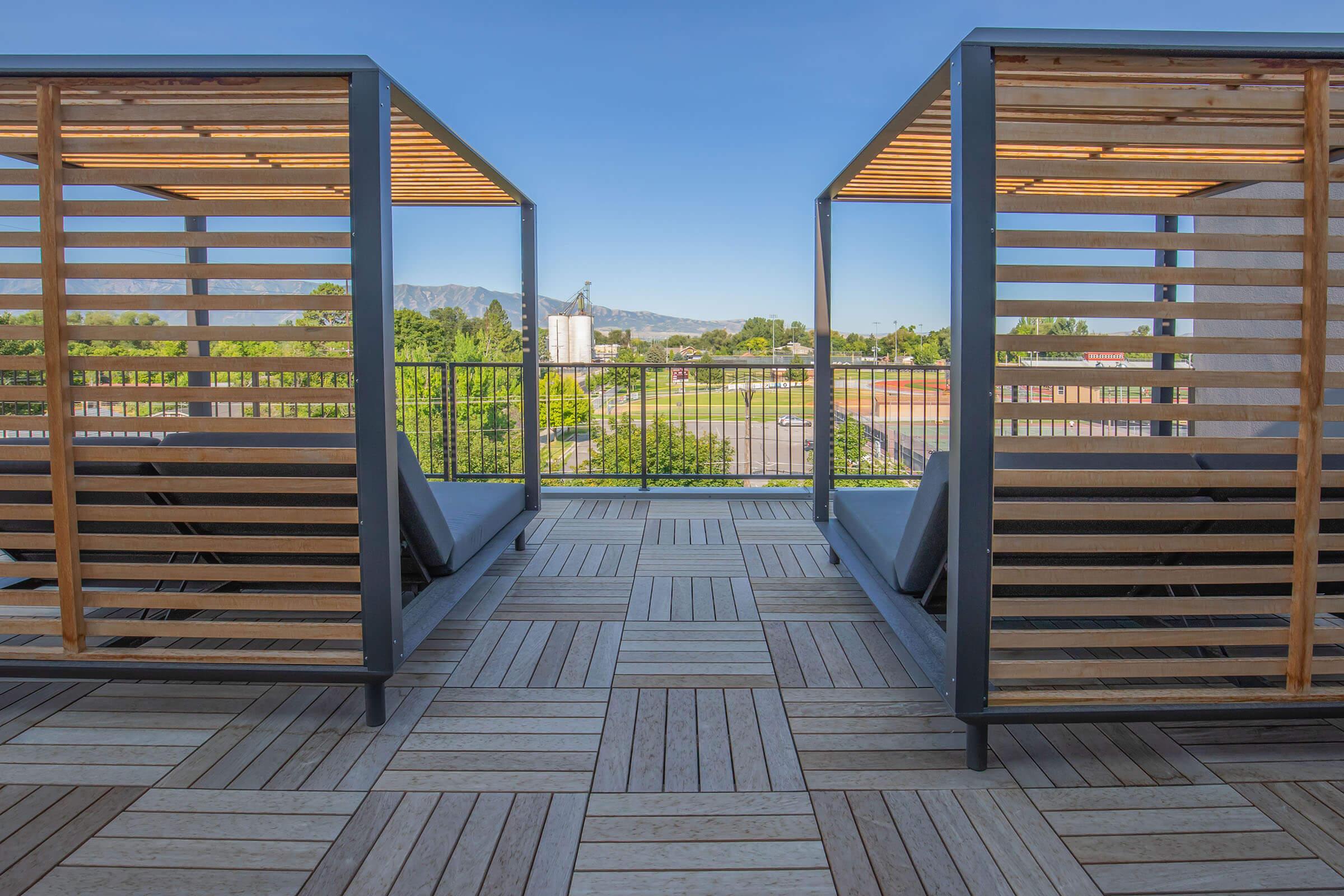 Two modern lounge areas with wood and metal frames, positioned on a rooftop terrace. Each area features a slatted design for privacy, with comfortable loungers facing a scenic view of green fields and distant mountains under a clear blue sky. The flooring consists of wooden planks arranged in a geometric pattern.