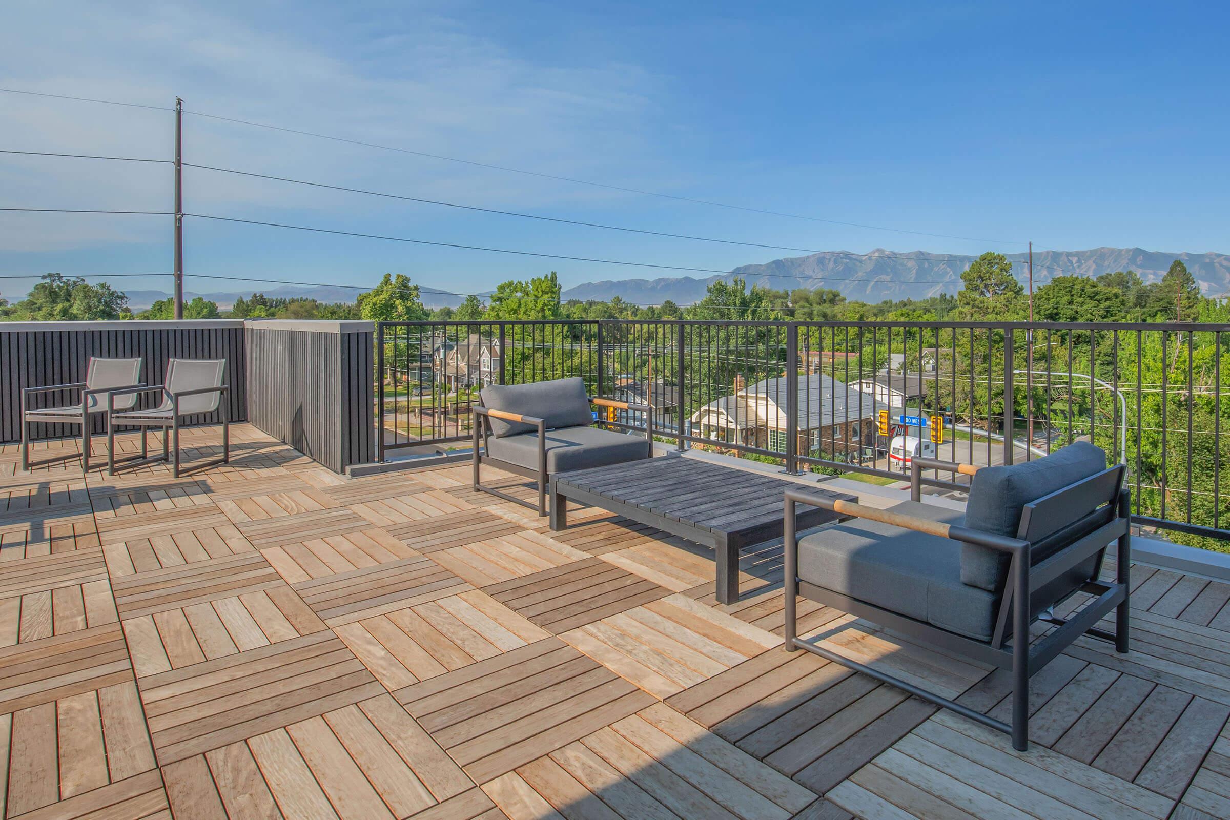 A rooftop terrace featuring modern outdoor furniture, including two seating areas with gray sofas and a dark wooden coffee table. The wooden decking creates a warm ambiance, and in the background, there are trees and mountains against a clear blue sky. Power lines are visible in the distance.