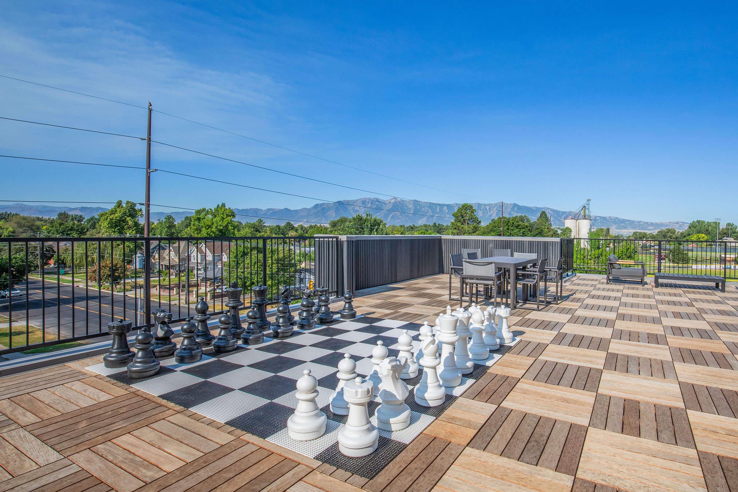 A rooftop terrace featuring a giant outdoor chess set with black and white pieces arranged on a large checkerboard. In the background, there are mountains under a clear blue sky, and the terrace is surrounded by green trees and a modern fence.