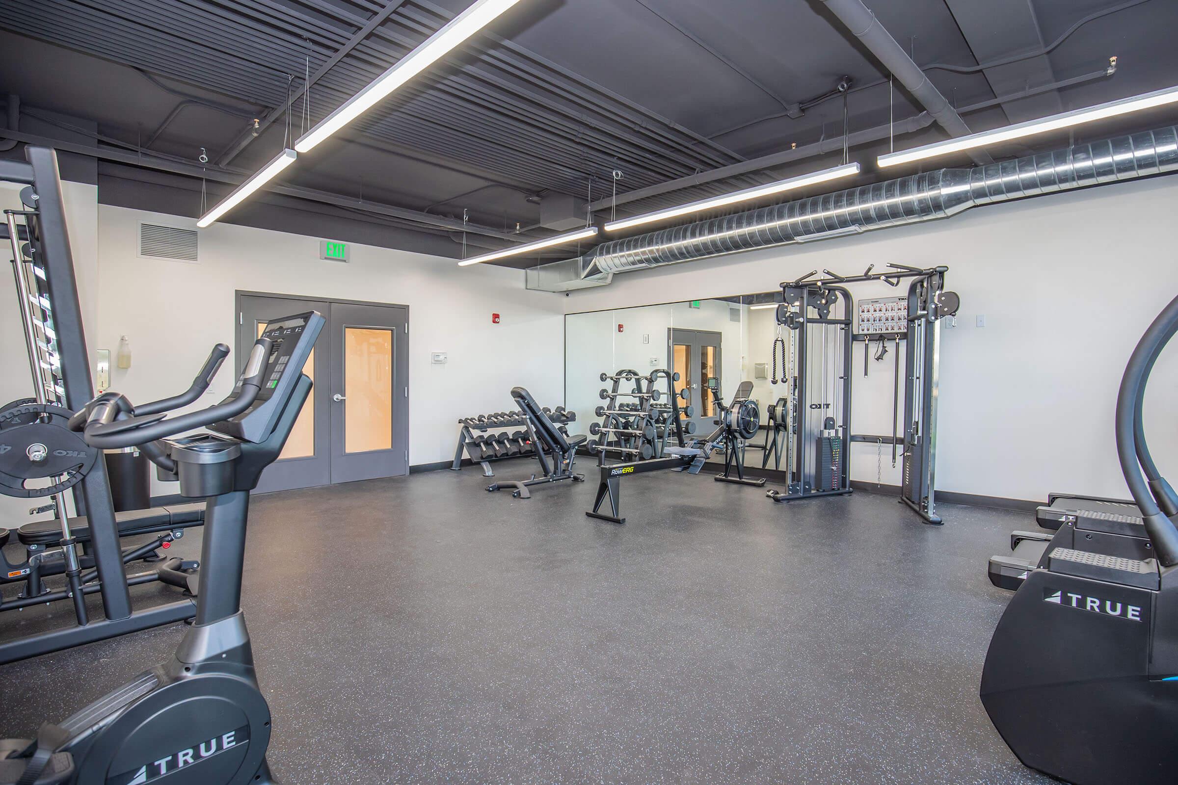 A clean and modern fitness room featuring various exercise equipment, including ellipticals, strength-training machines, and free weights. Large mirrors line one wall, and the space has a polished floor and bright overhead lighting. The atmosphere is inviting for workouts and fitness activities.