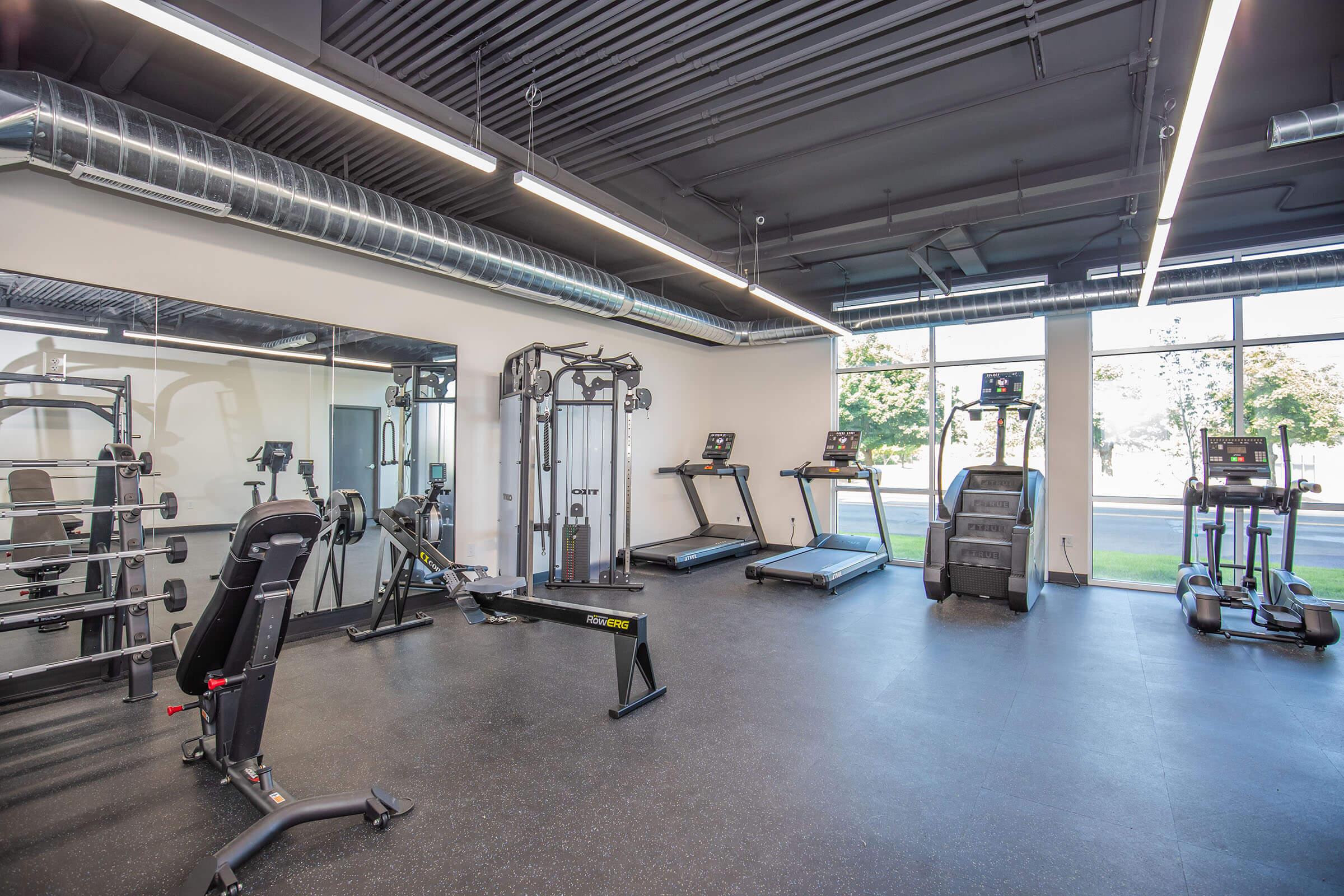 A modern gym interior featuring various exercise equipment. The space includes treadmills, a weight machine, a bench press, and free weights. Large windows allow natural light to fill the room, creating a bright and inviting atmosphere for workouts. The flooring is covered with dark rubber mats for safety.