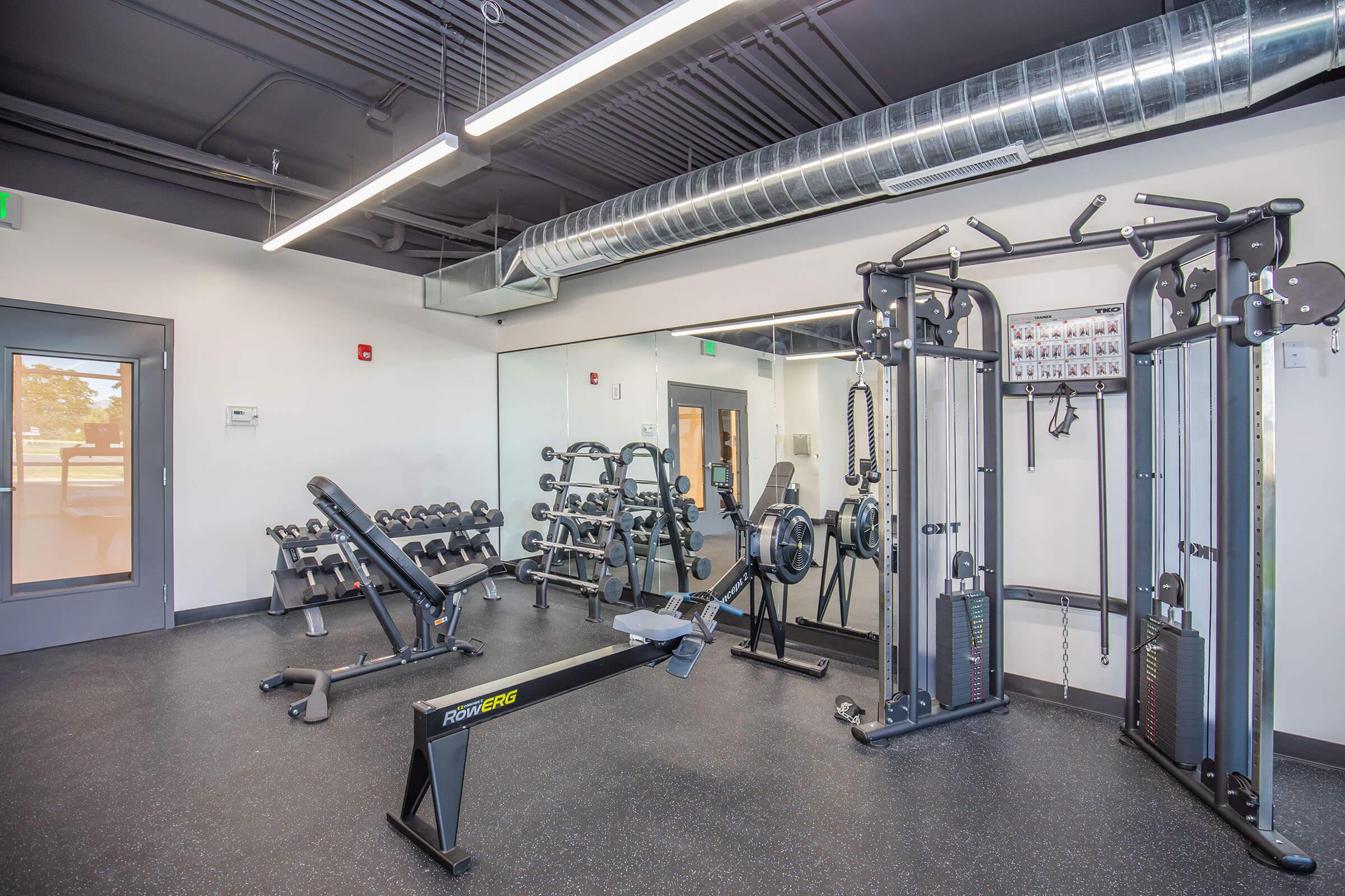 A modern gym interior featuring free weights, a cable machine, and a multi-purpose bench. The space has large mirrors on one wall, black rubber flooring, and bright overhead lighting, creating an inviting atmosphere for workouts.
