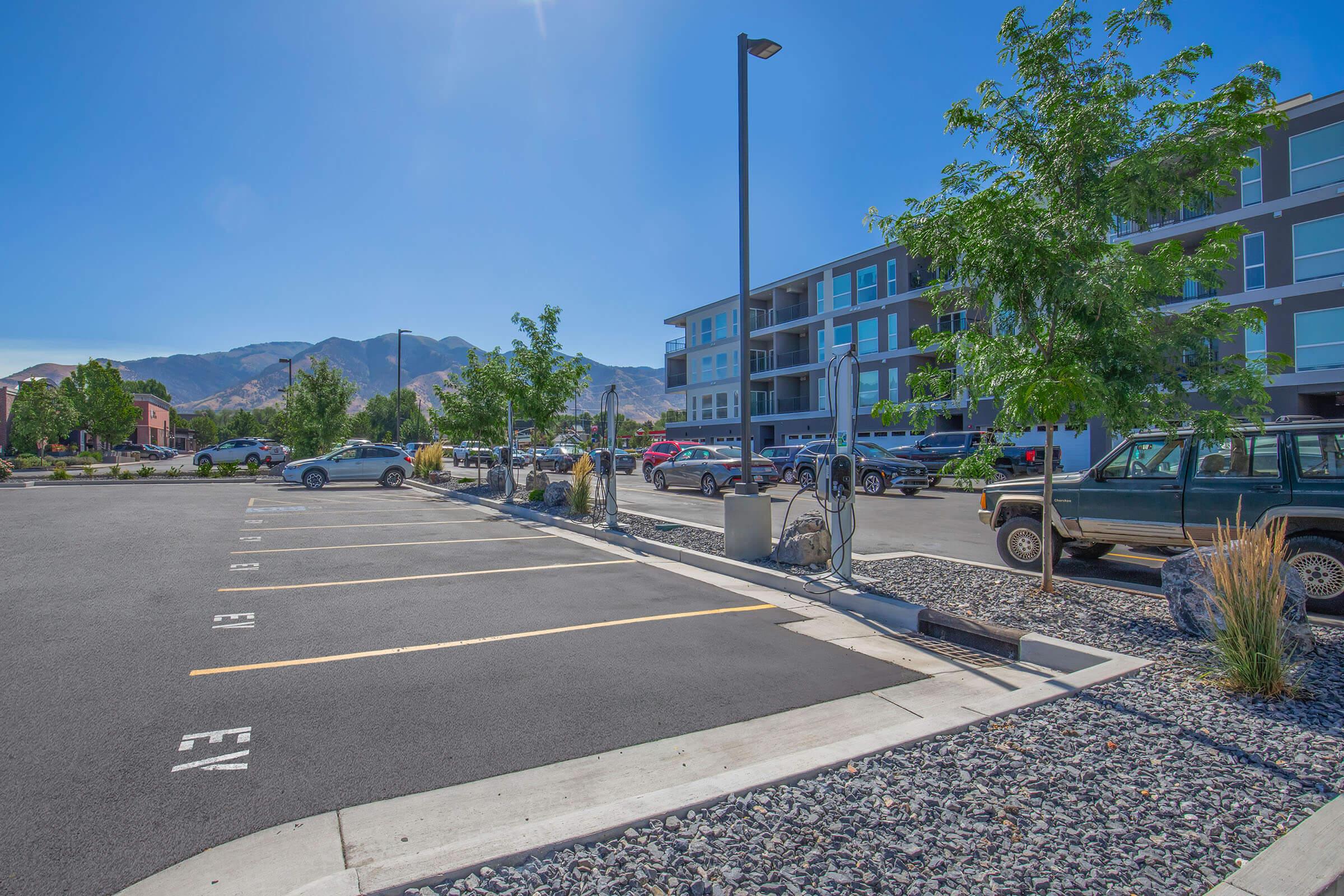 A parking lot with empty spaces, electric vehicle charging stations, and a few vehicles. In the background, there are modern apartment buildings and mountains visible under a clear blue sky. Trees and landscaping include rocks and shrubs, adding greenery to the area.