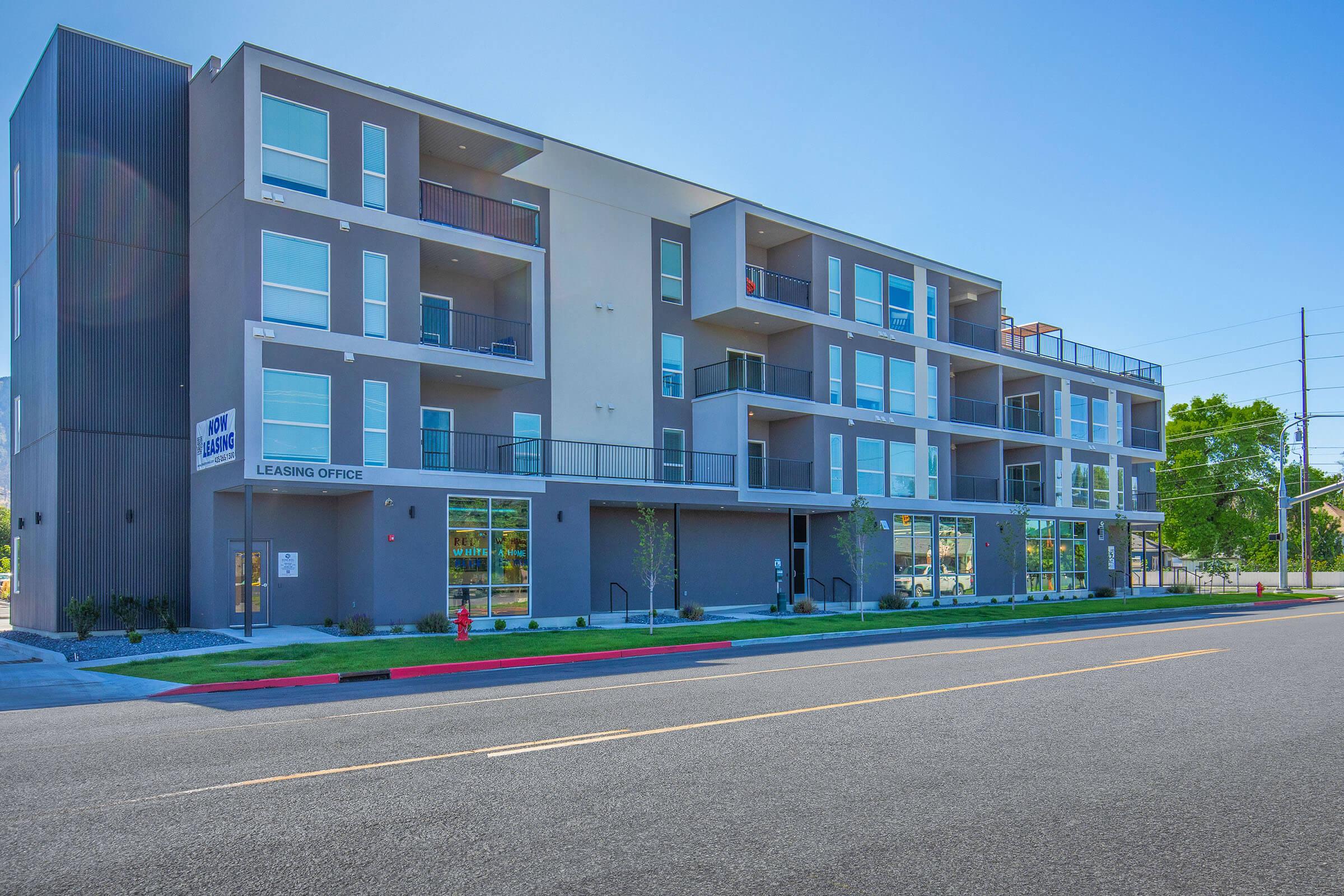 Modern multi-story apartment building with a leasing office on the ground floor. The façade features a mix of gray and white materials, large windows, and several balconies. The building is situated on a street with clear blue skies and greenery nearby.