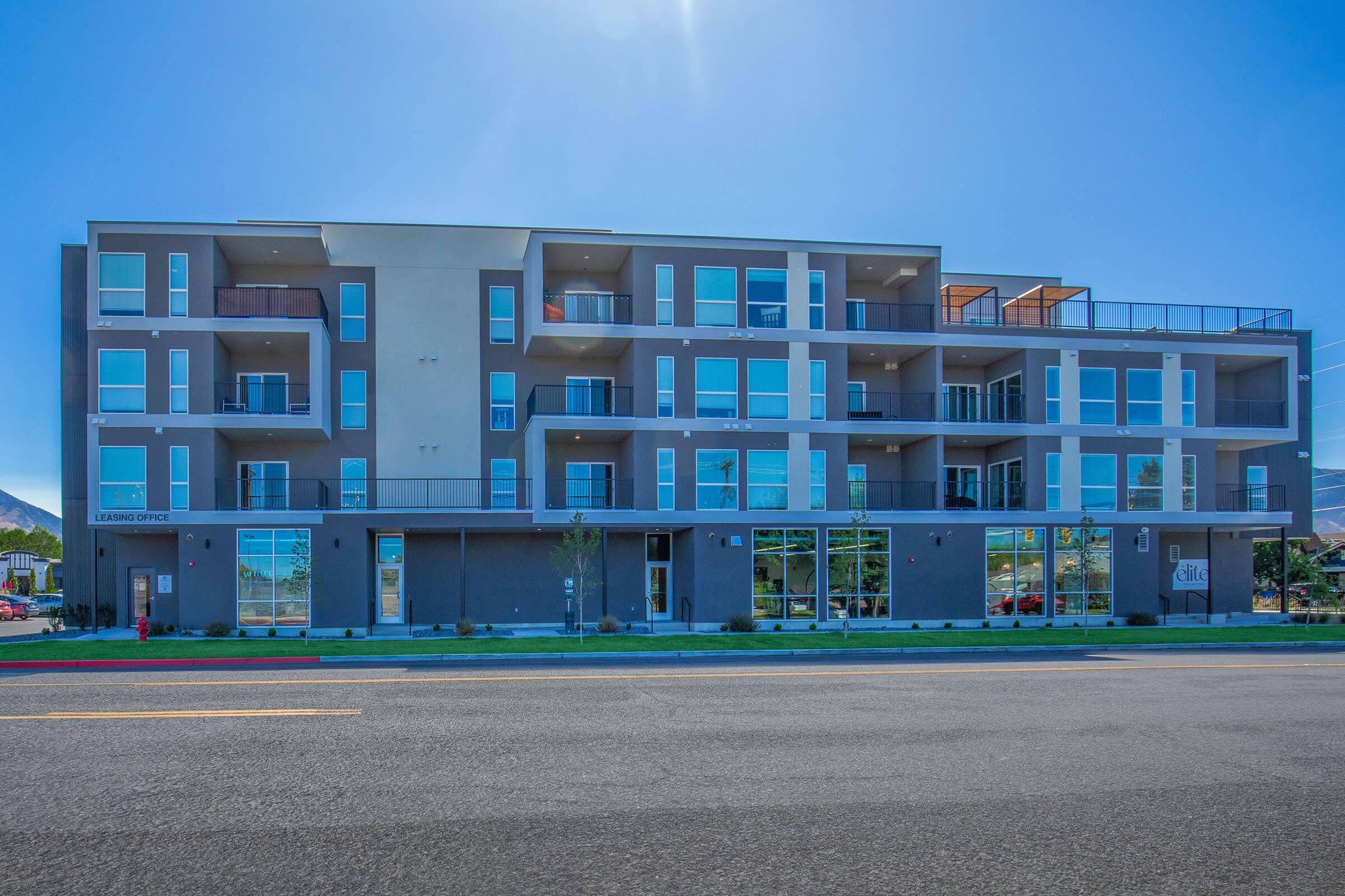 Modern multi-story apartment building with a sleek design, featuring several balconies and large windows. The ground floor includes a leasing office with glass doors, surrounded by landscaped areas and a clear blue sky. A road runs in front of the building, indicating a bustling urban setting.