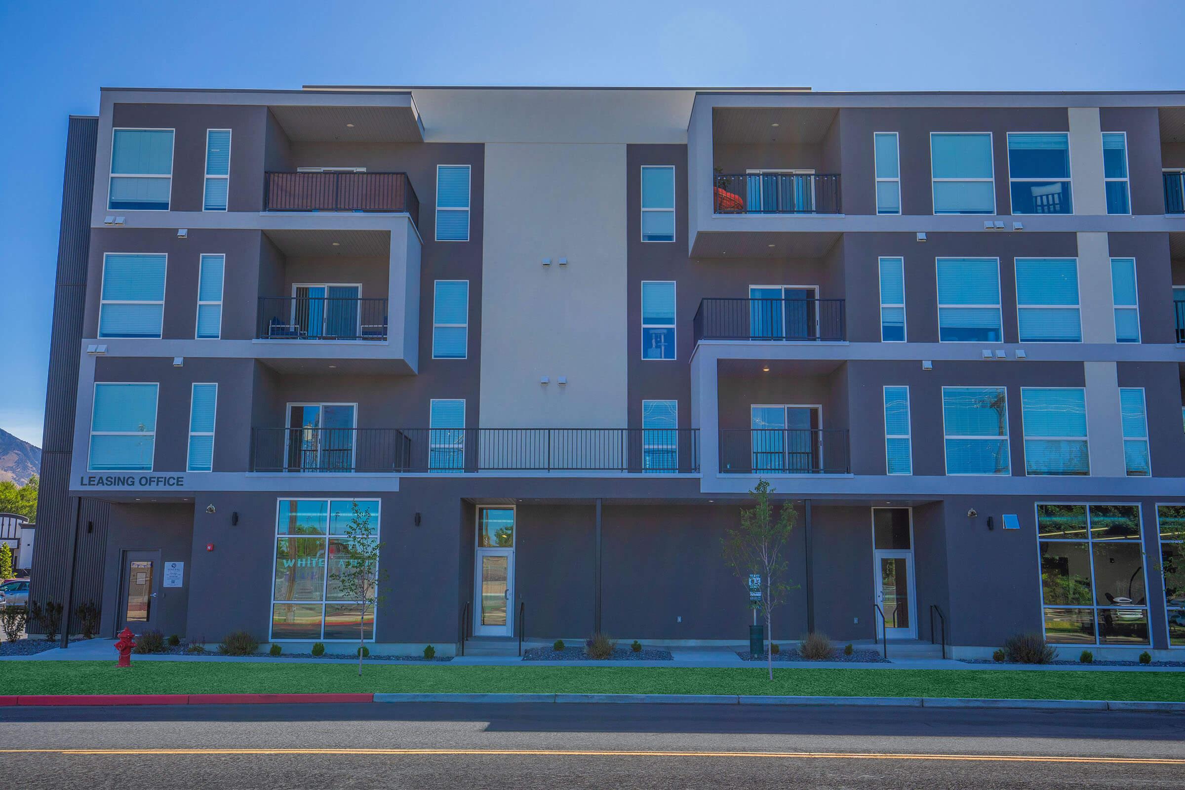 Exterior view of a modern multi-story apartment building with a leasing office. The structure features a mix of large windows and balconies, and is set against a clear blue sky. The ground area is landscaped with small shrubs and grass, and there is a fire hydrant visible near the sidewalk.