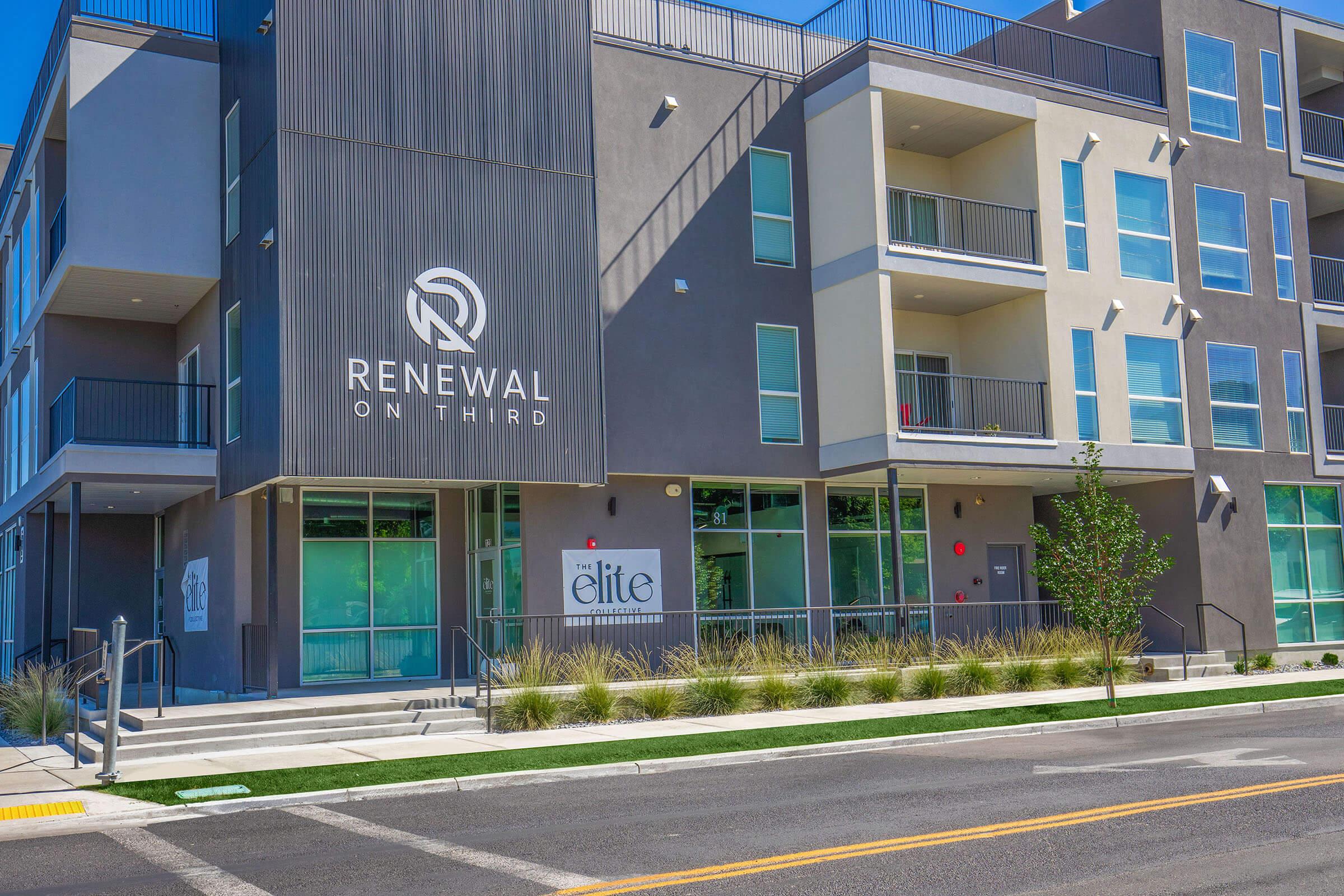 Modern apartment building with a sleek facade featuring large windows and balconies. The sign "Renewal on Third" is prominently displayed. A smaller sign indicates "The Elite" at the entrance. The surrounding landscape includes neatly trimmed grass and shrubs along the sidewalk.