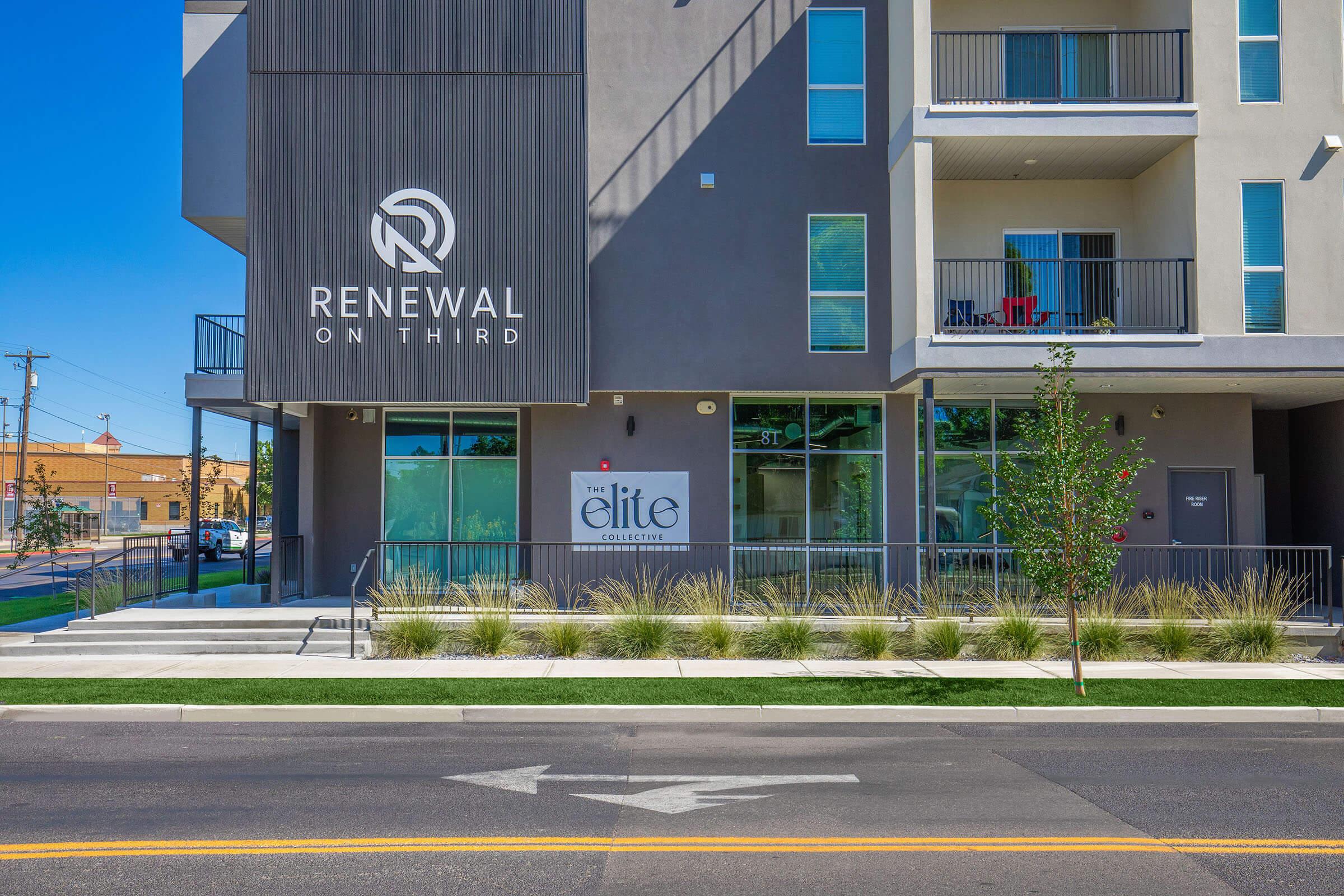 A modern multi-story building with a sign that reads "Renewal on Third" in a sleek font. The ground level features large windows and an entrance with a sign for "The Elite Collective." The exterior is a combination of gray and white, surrounded by well-maintained landscaping and a clear blue sky.
