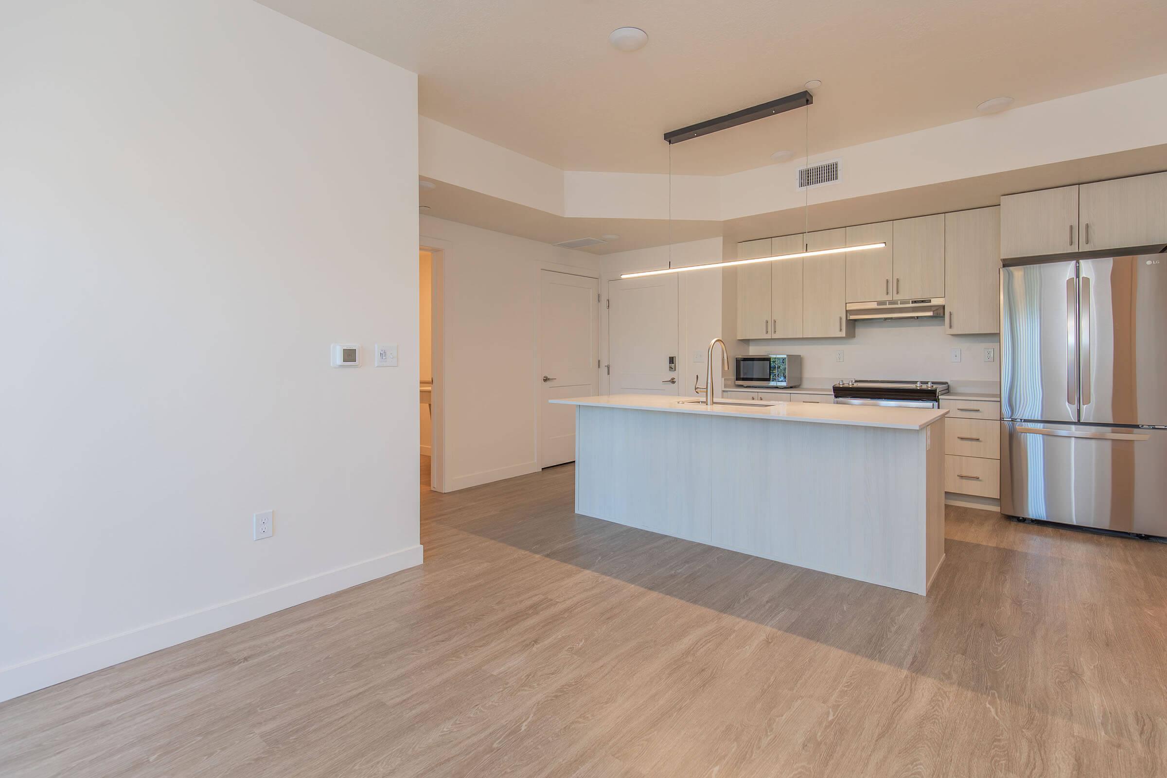 Modern kitchen and living area in an apartment featuring light wood flooring, a kitchen island with a sink, stainless steel appliances, and ample cabinetry. The space is well-lit with overhead lighting, and there is an open layout leading to another room.
