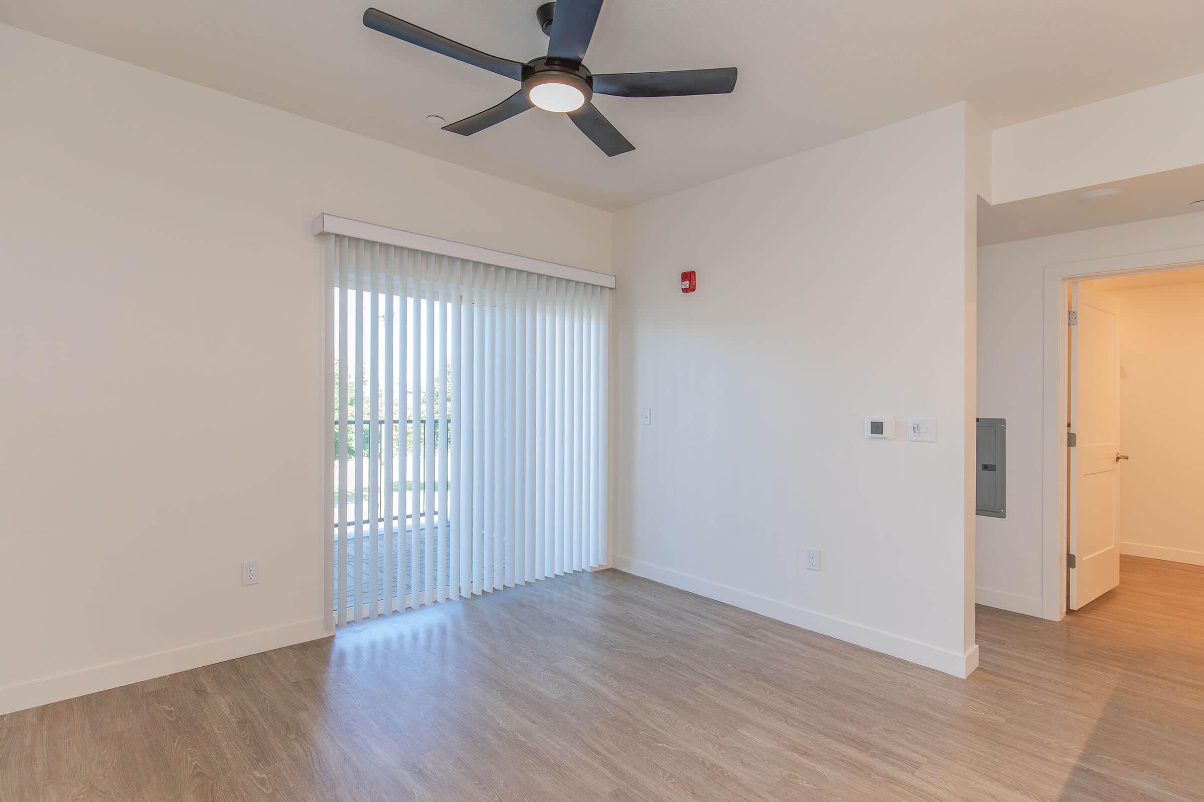 Bright and spacious interior of a modern living room featuring a ceiling fan, large window with vertical blinds, and light-colored walls. The flooring is hardwood, and there is a doorway leading to another room. The overall ambiance is airy and welcoming.