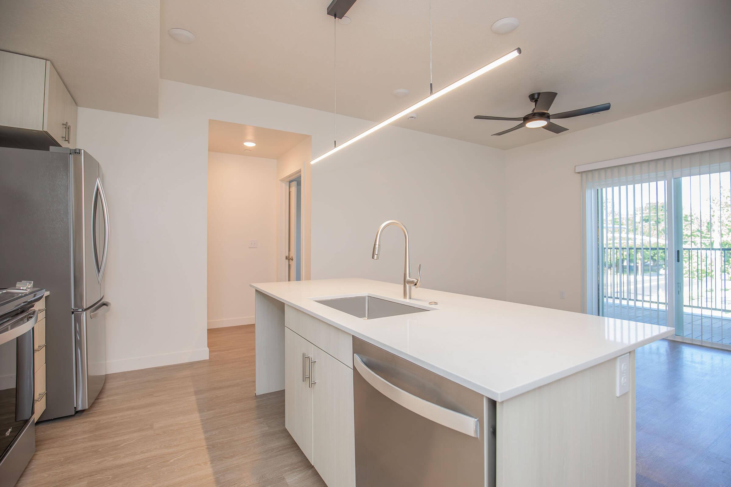 Modern kitchen with an island sink, stainless steel appliances, and a sleek design. The room features white walls, wood flooring, and a large window with sliding doors leading outside. Bright, minimalist lighting hangs above the countertop, enhancing the spacious feel.