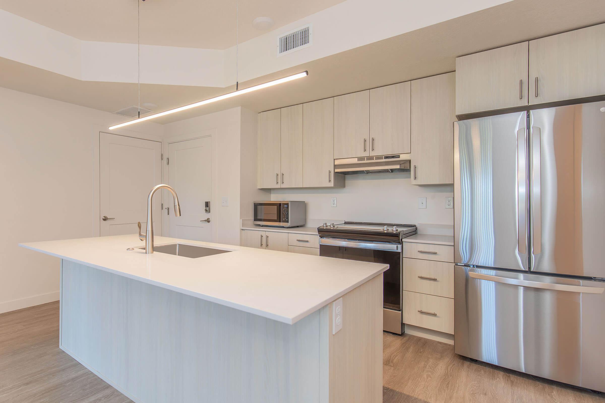 Modern kitchen interior featuring light wood cabinetry, a large island with a sink, stainless steel appliances including a refrigerator and oven, and a microwave. The space has a bright, airy feel with neutral colors, and a contemporary lighting fixture hanging above the island.
