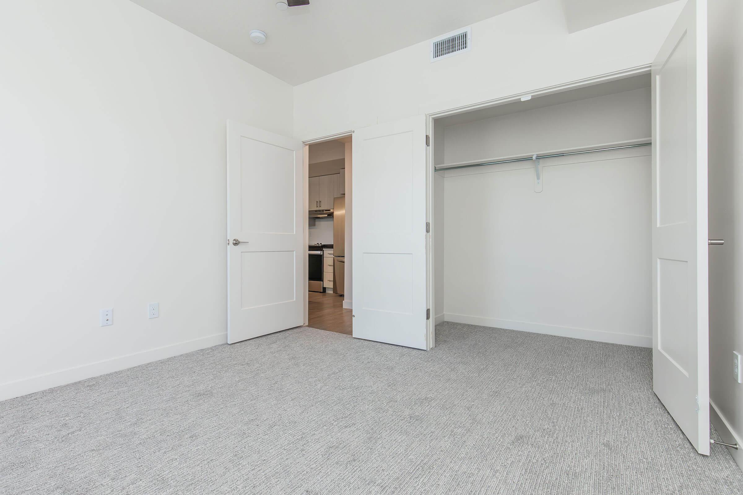 A spacious room with light-colored walls and carpeted flooring. Two white doors lead to a closet and another area. The closet has a hanging rod and is empty. The room is well-lit, suggesting a modern design, with a glimpse of a kitchen area visible through an open door.