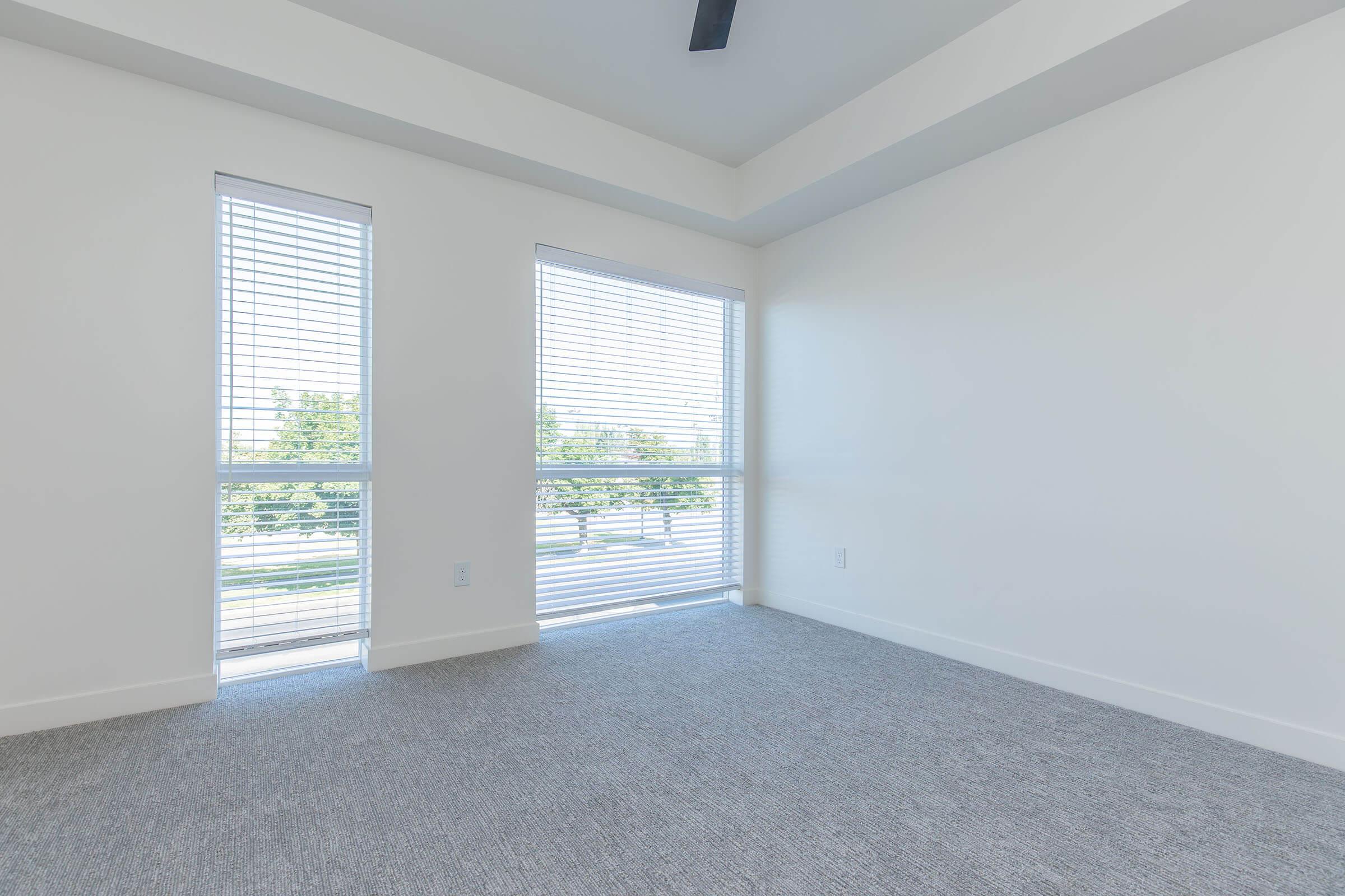 A bright, empty room with large windows featuring blinds and light-colored walls. The floor is covered with grey carpet, and there is a ceiling fan visible. The space is minimalistic, offering natural light and a view of greenery outside.