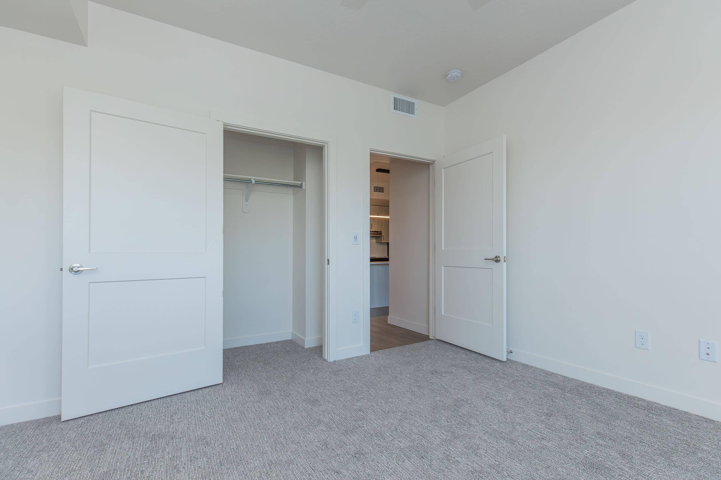 A spacious room featuring light-colored walls and carpet, with two open doors leading to closets and another area. Natural light illuminates the space, highlighting its clean, modern design.