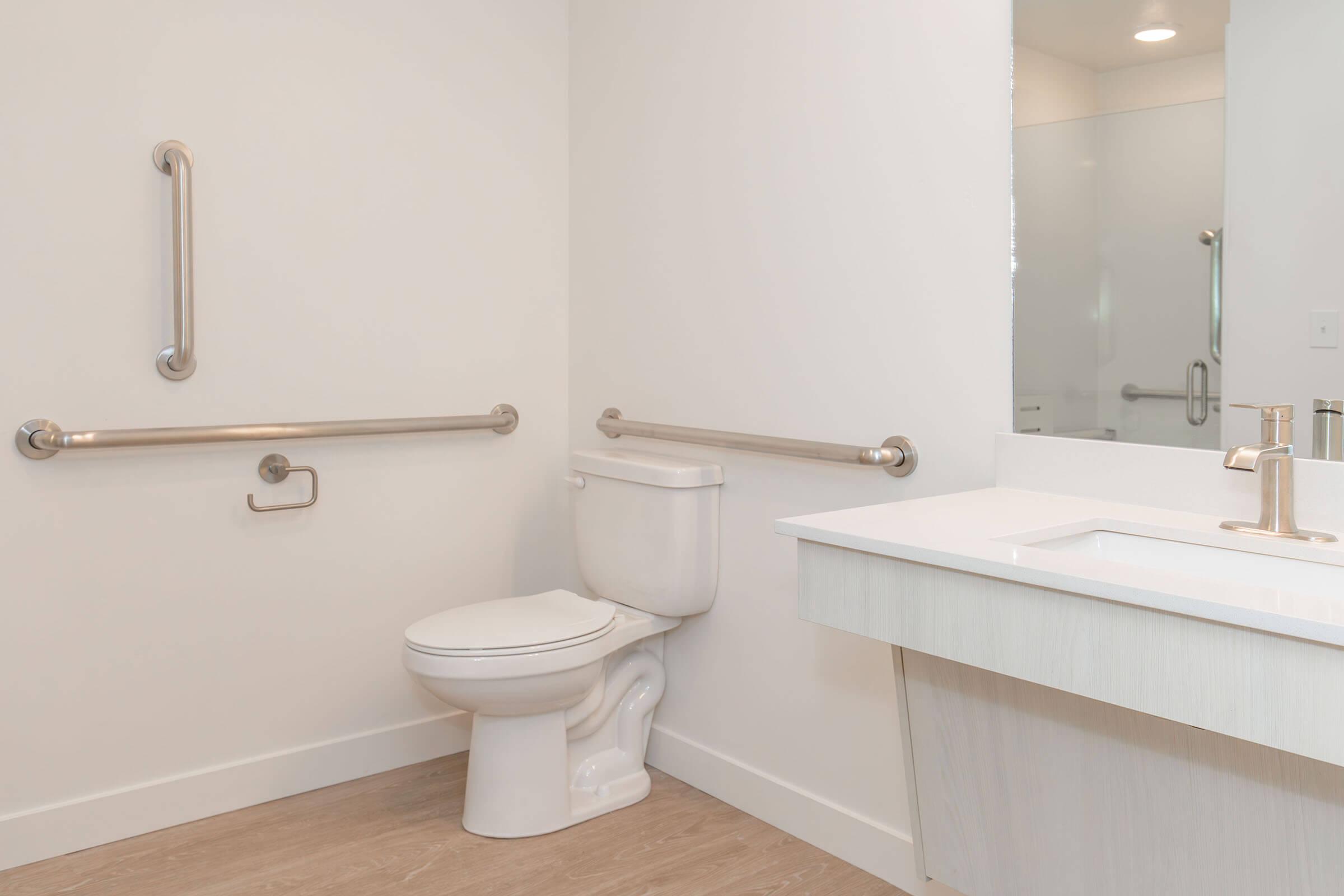 A clean, modern restroom featuring a white toilet, grab bars for accessibility, and a simple sink with a mirror. The walls are painted in a light color, and the flooring is a wood-like material, promoting a bright and airy ambiance.