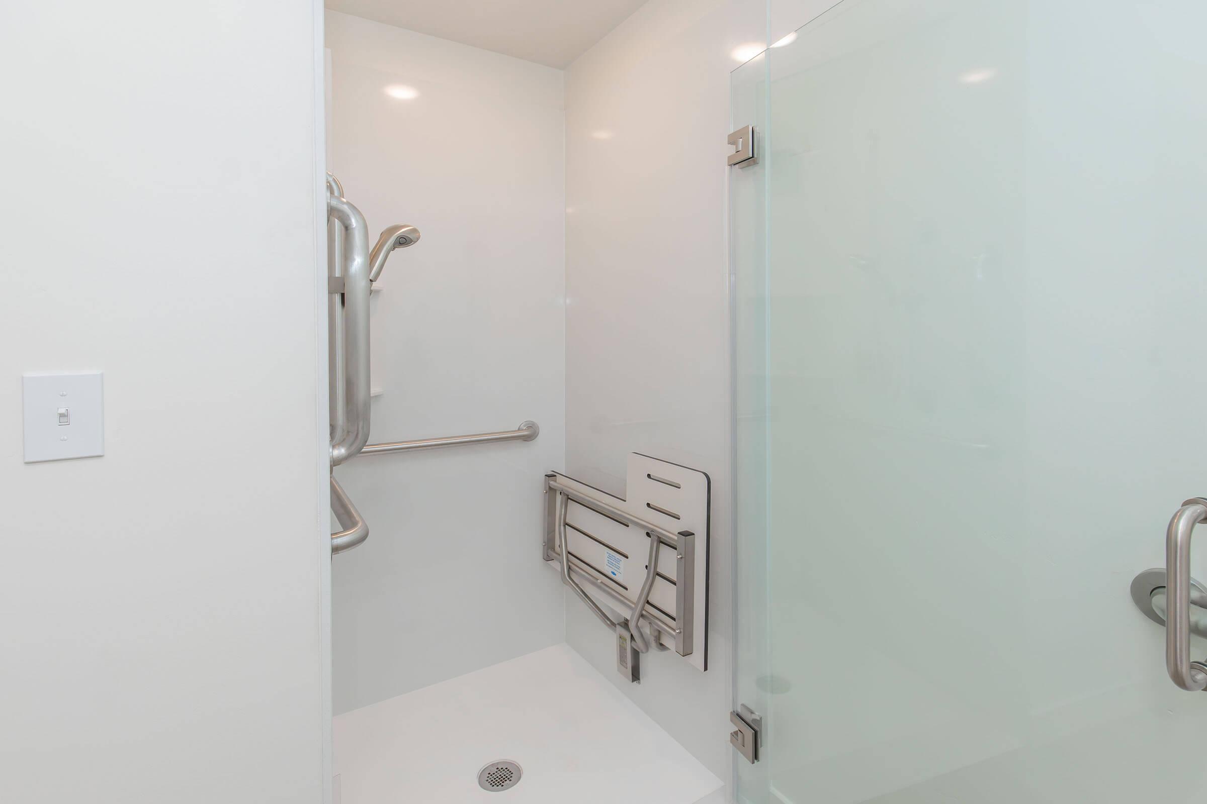 A modern, accessible shower with a glass door, a foldable seating area, and grab bars for safety. The walls are light-colored, providing a clean and bright appearance. The floor is tiled and features a drain at the center.