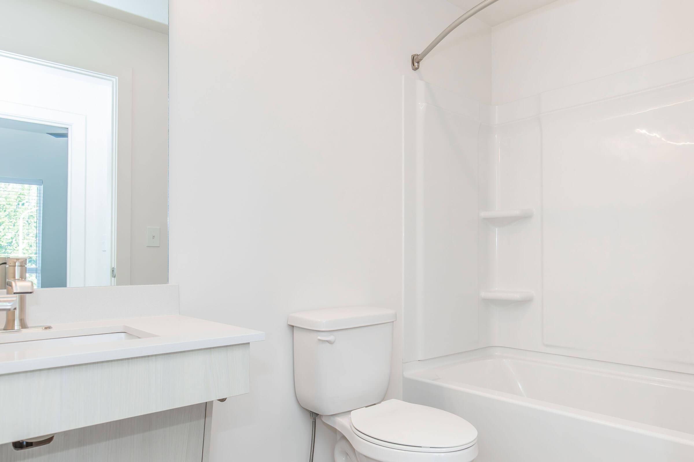 A clean, modern bathroom featuring a white bathtub and shower combo, a white toilet, and a sleek vanity with a sink. The walls are light-colored, and a mirror is visible in the background, reflecting a light and airy atmosphere. Natural light is coming in from an unseen window.