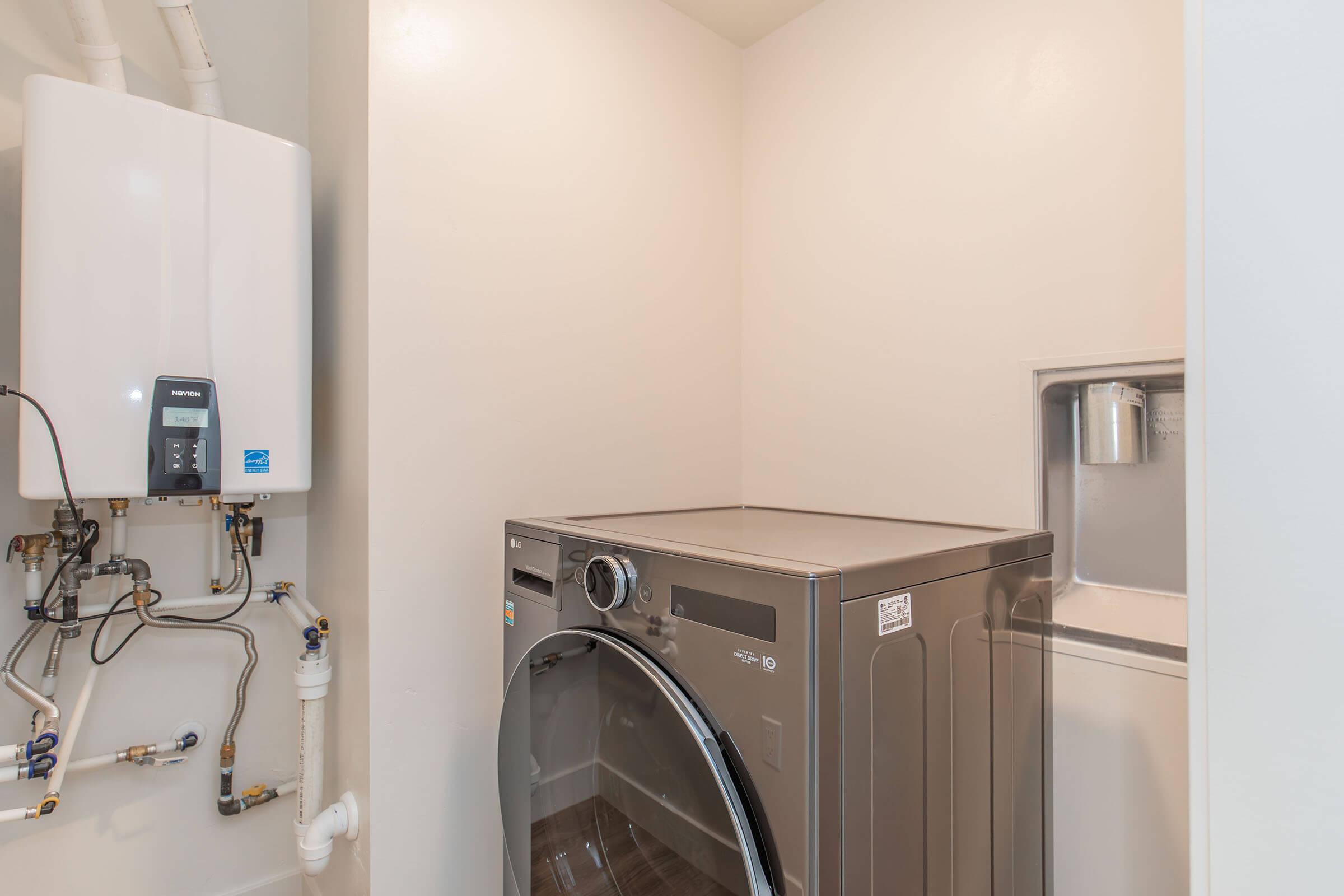 A laundry room featuring a modern washing machine beside a compact water heater mounted on the wall. The walls are painted white, and the space is well-lit, highlighting the appliances and the clean, organized environment. There are plumbing connections visible near the heater.
