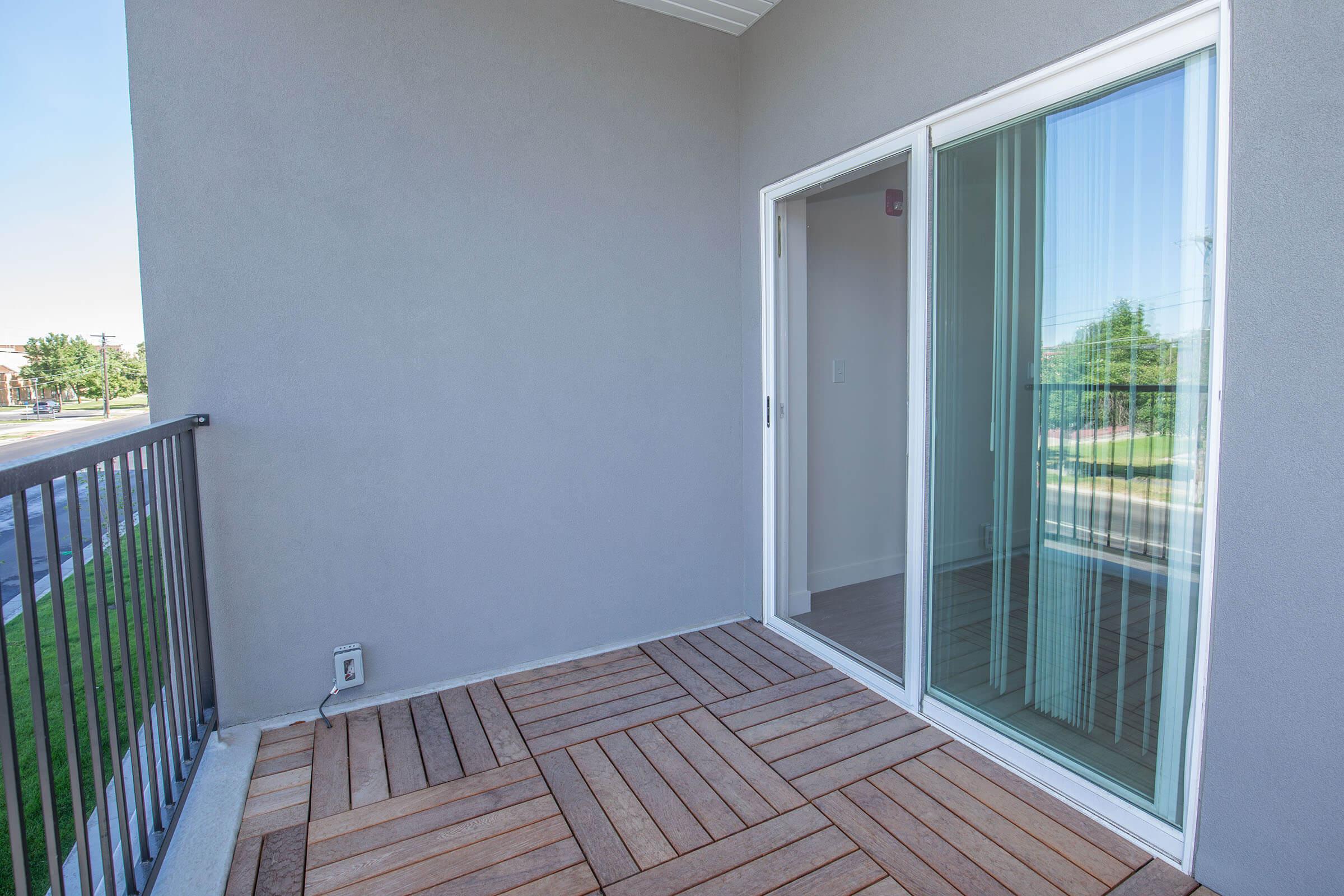 Empty balcony with wooden flooring and a glass sliding door, leading to an indoor space. A grey exterior wall is visible, and outside there are hints of greenery and structures beyond the railing. The area is well-lit, suggesting a bright day.