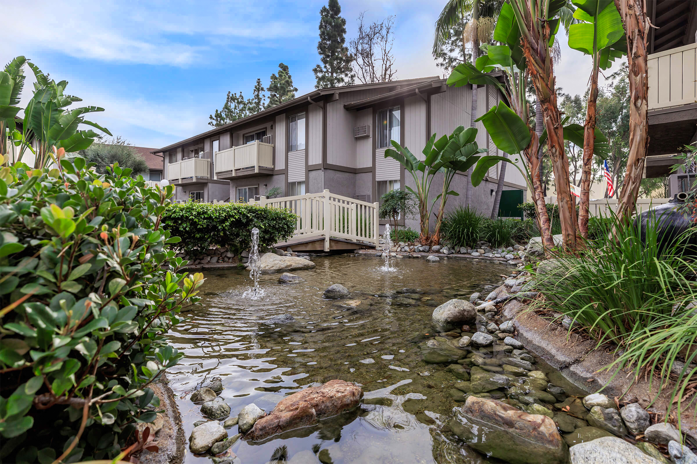 A tranquil courtyard scene featuring a small pond with gentle water fountains, surrounded by lush greenery and rocks. In the background, a two-story apartment building with balconies and tropical plants adds to the serene atmosphere. The sky is clear and bright, enhancing the peaceful setting.