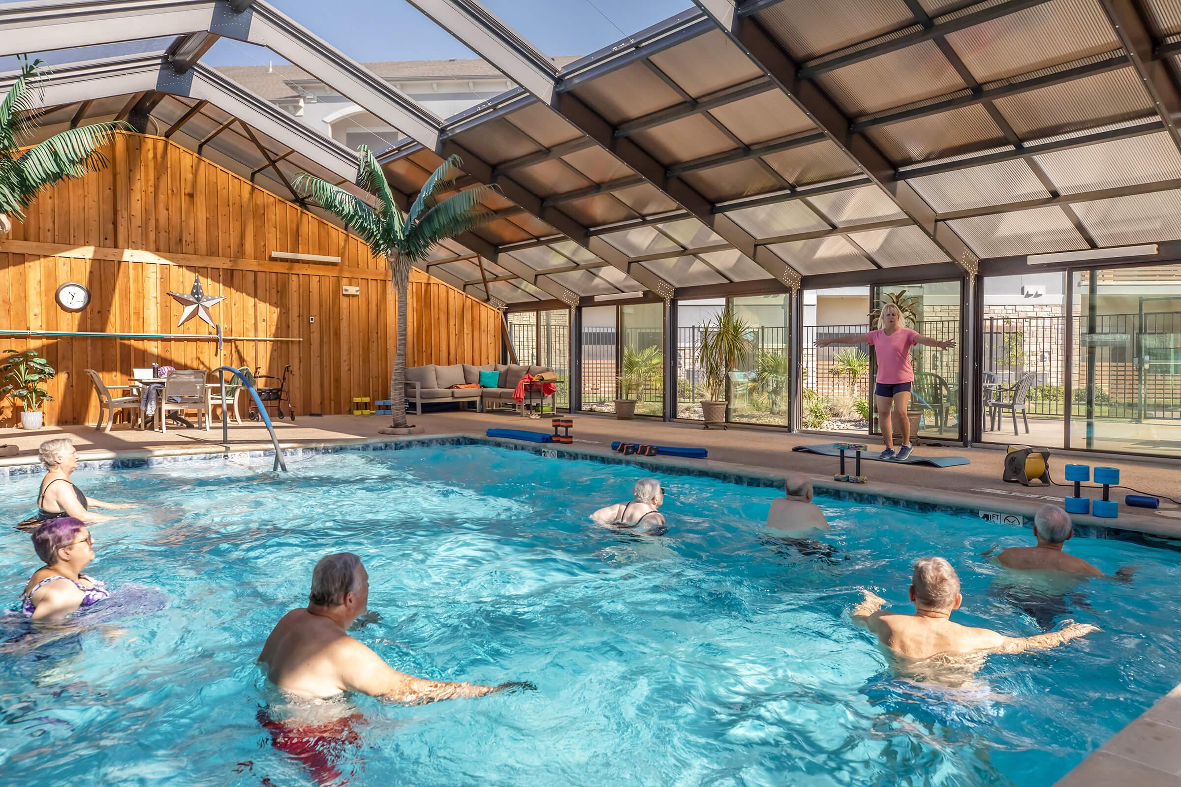 An indoor swimming pool area with large windows allowing natural light. Several seniors are participating in a water aerobics class led by an instructor at the edge of the pool. The space features wooden paneling, potted plants, and exercise equipment around the pool.