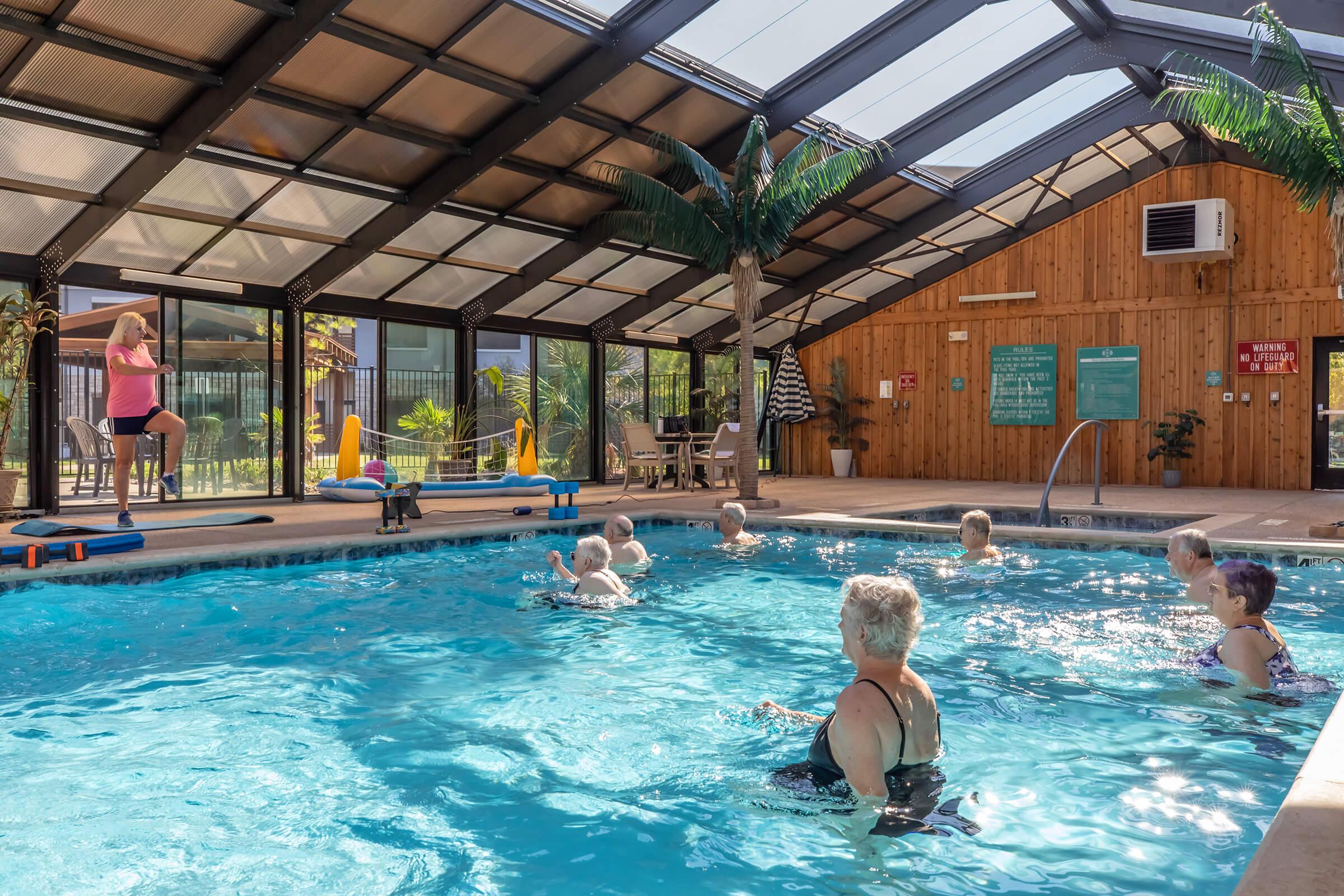 A bright indoor pool area with a clear ceiling, featuring several people participating in a water aerobics class. Some individuals are exercising in the pool while others watch. Palm trees decorate the space, adding a tropical feel, and there are lounge chairs and poolside signs visible in the background.