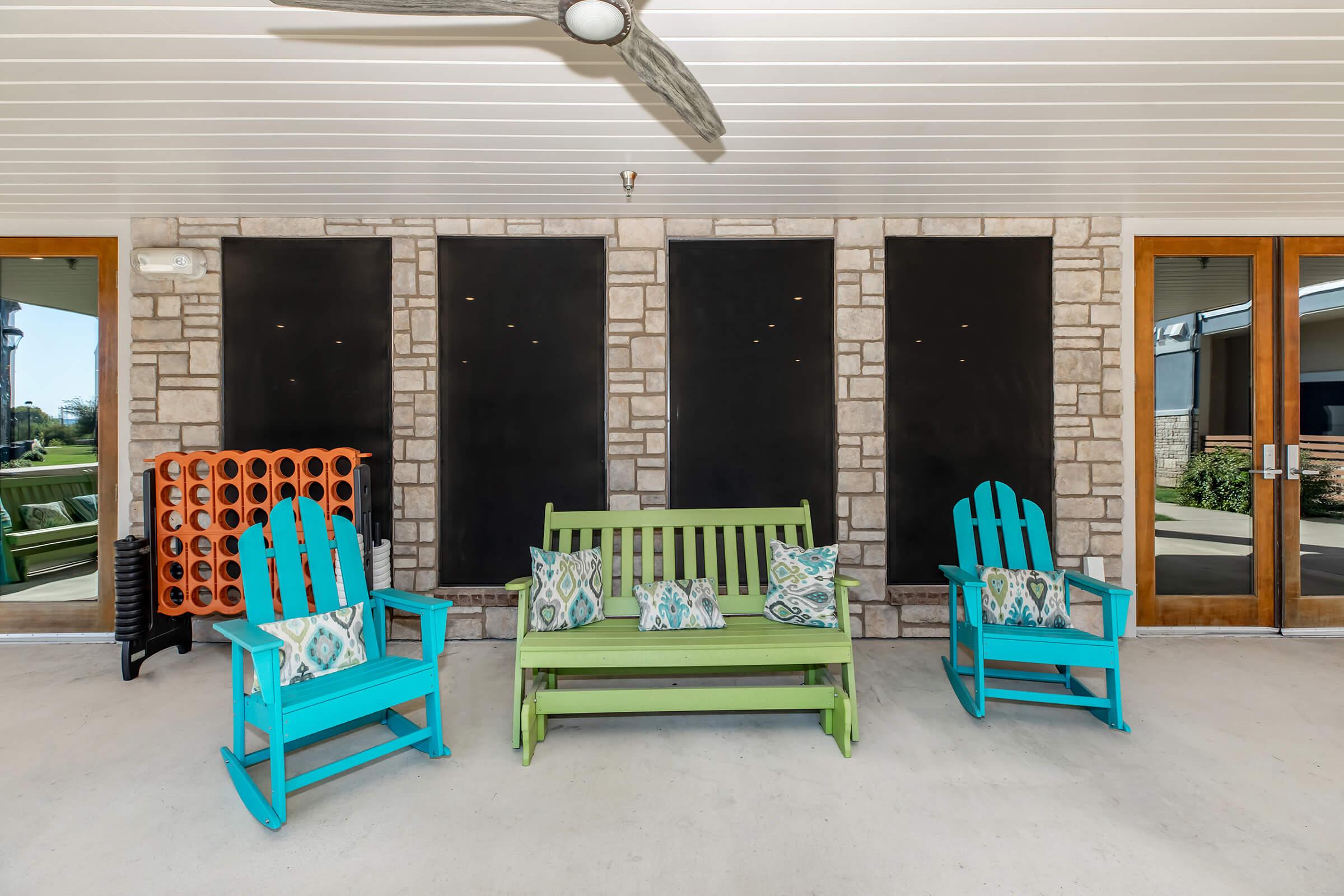 A cozy outdoor patio featuring a green wooden bench and two bright blue Adirondack chairs with decorative pillows. The background has a stone wall with dark panels, and there's a rack with orange and black circular storage. A ceiling fan is visible above, enhancing the inviting atmosphere.