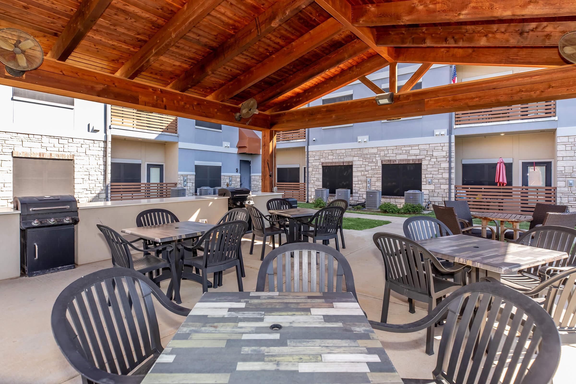 Outdoor seating area under a wooden gazebo, featuring several black chairs and tables. A barbecue grill is positioned to one side, while the gazebo's structure provides shade. The backdrop consists of a modern building with patio doors and grass landscaping, creating a welcoming atmosphere for gatherings.