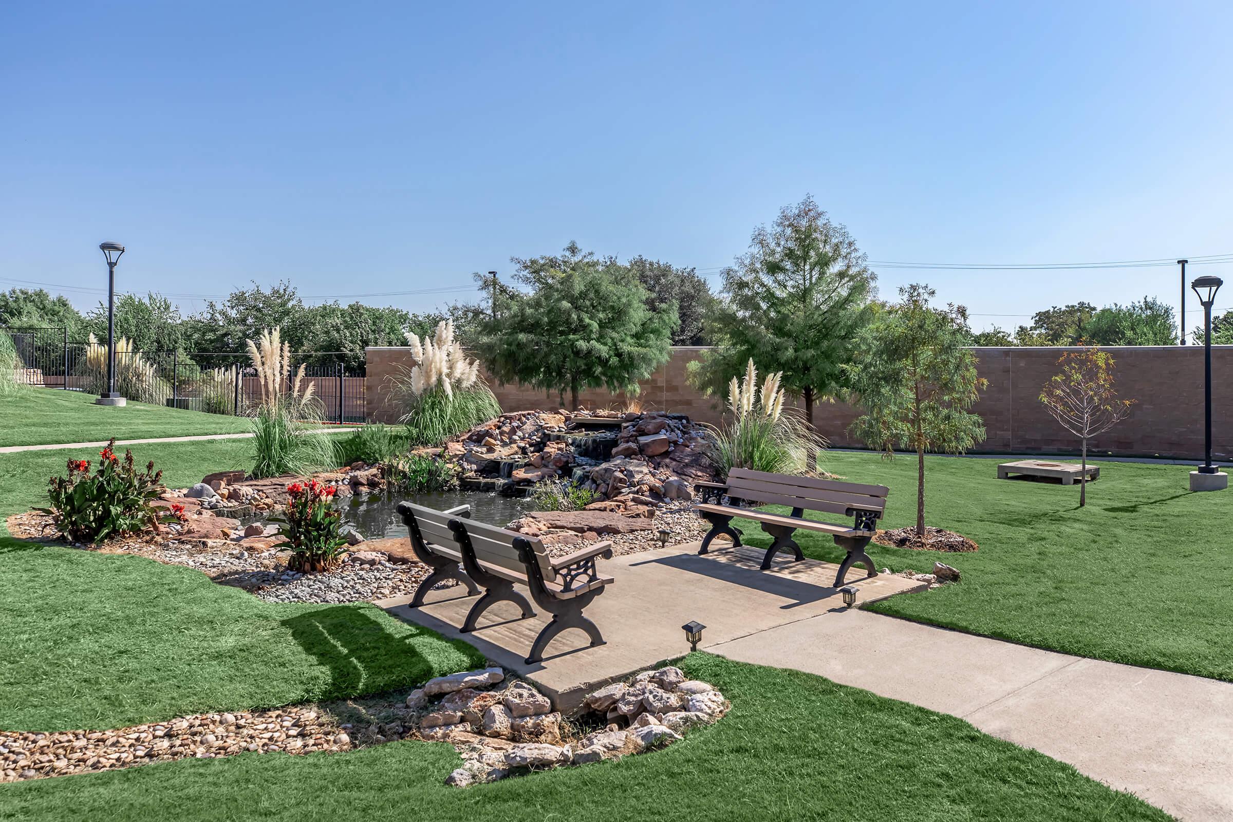 A serene outdoor garden featuring a small pond with a decorative rock waterfall, surrounded by greenery and flowering plants. There are two wooden benches positioned on a paved path, with additional landscaping including ornamental grasses and trees. Clear blue sky overhead.