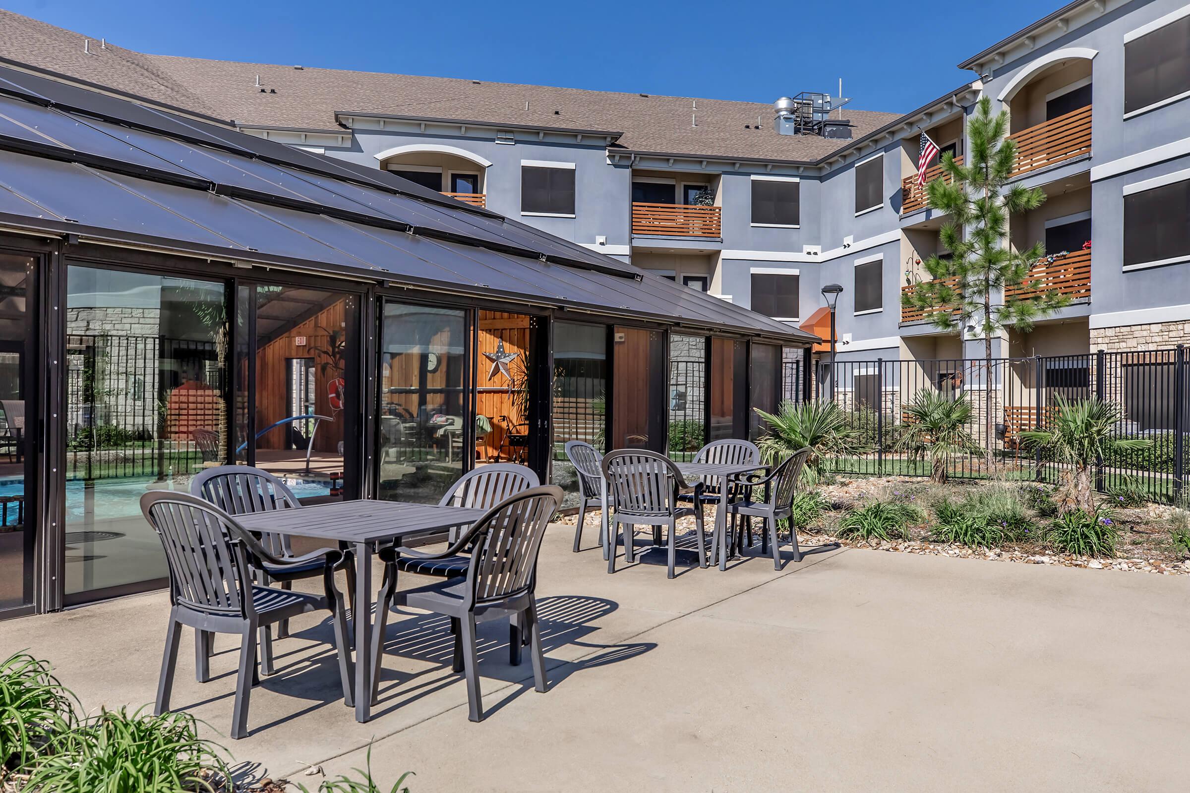 Outdoor seating area with several tables and chairs in front of a glass-enclosed patio. The background features a multi-story building with balconies and greenery. A clear blue sky is visible above, providing a bright and inviting atmosphere.