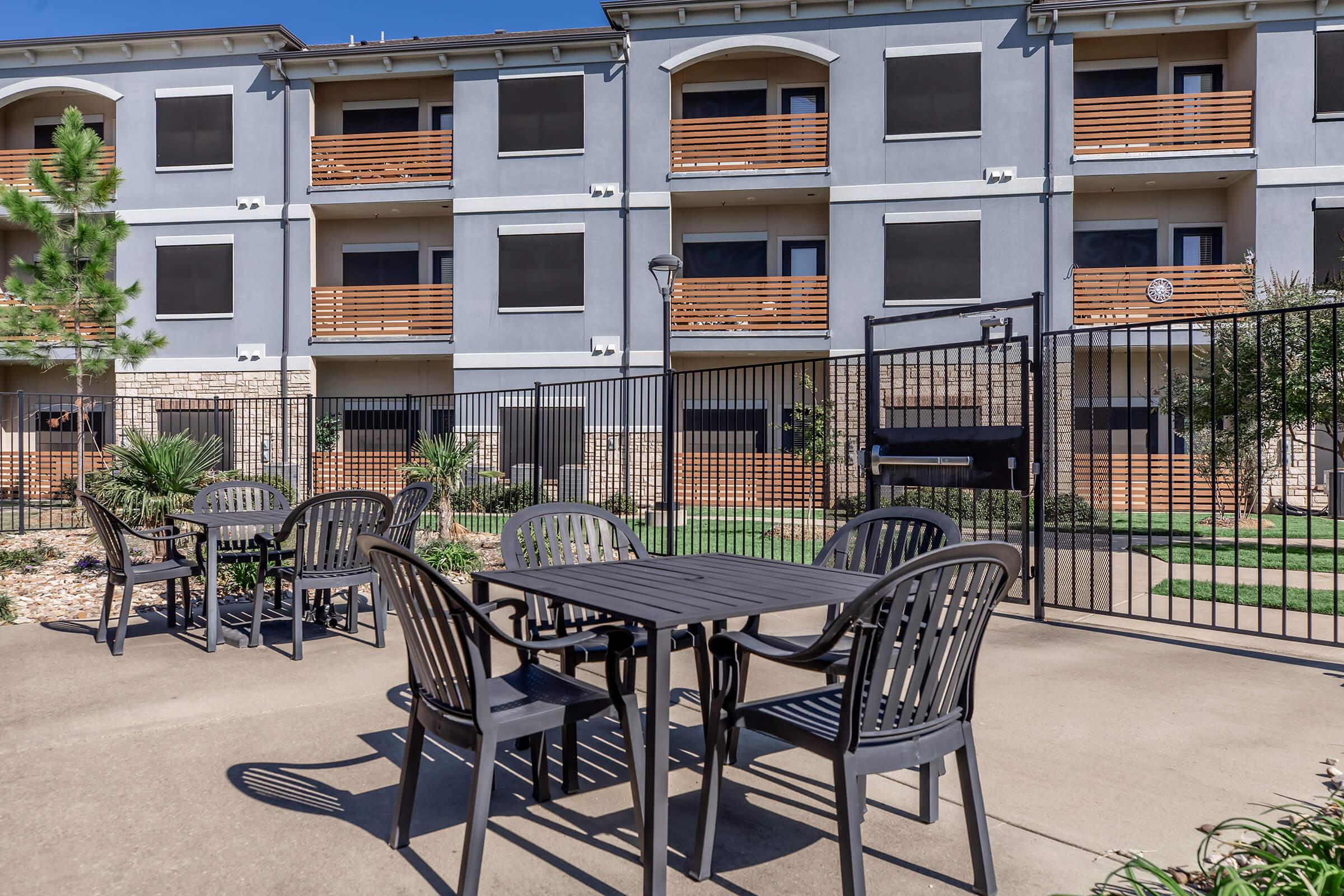 A view of an outdoor area with several black patio tables and chairs, surrounded by greenery. The background features a modern building with multiple floors, large window screens, and wooden balconies. A gate is visible, indicating an enclosed space.