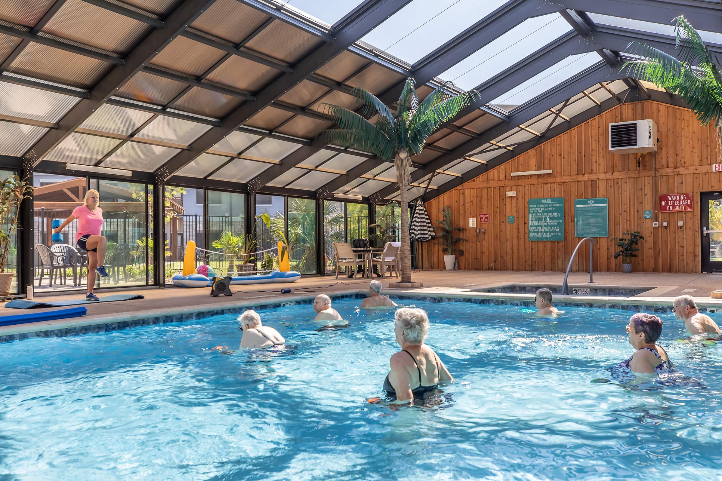 A group of elderly individuals participating in a water aerobics class in a bright indoor pool area. The space features large windows and a wooden wall, with palm tree decorations, as a female instructor leads the class. Floating devices are visible in the pool.
