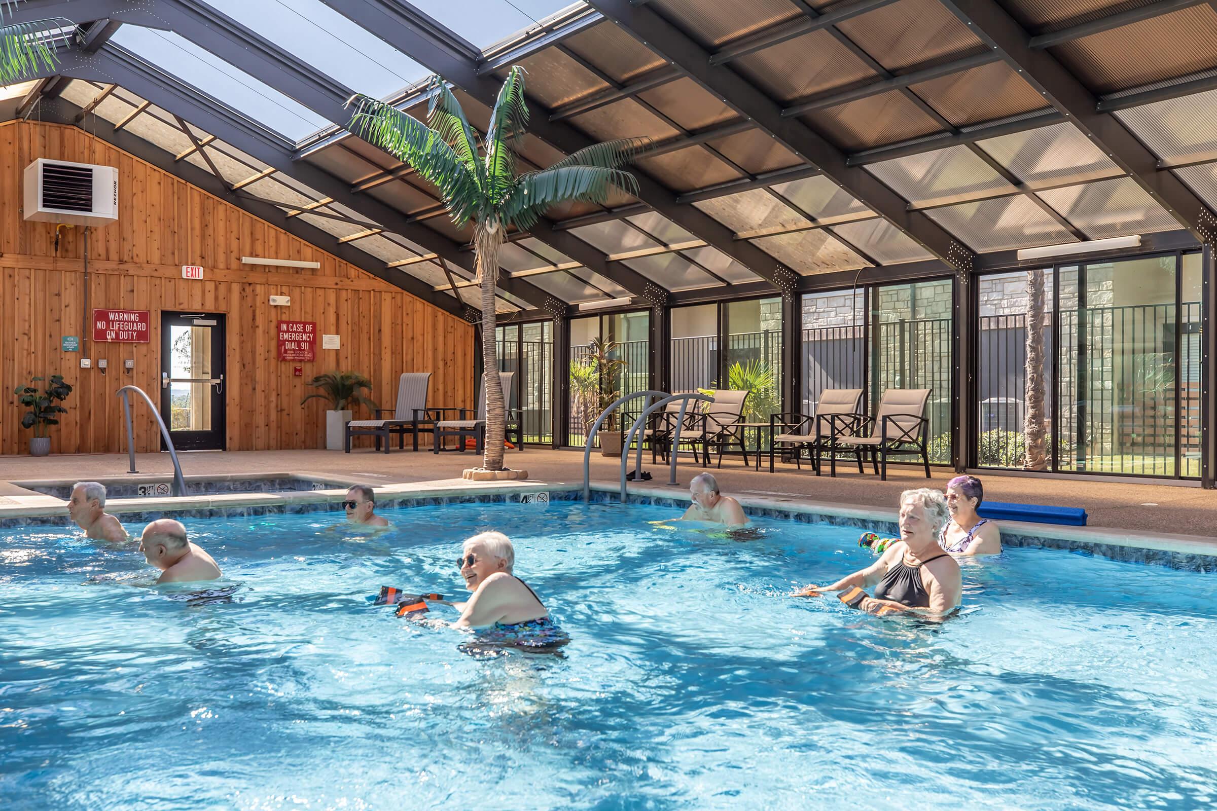 Indoor swimming pool with a glass roof, featuring several elderly individuals enjoying the water. They are engaged in various activities, with some using pool floats. The surrounding area includes lounges, potted plants, and walls made of wood and glass, creating a relaxing atmosphere.