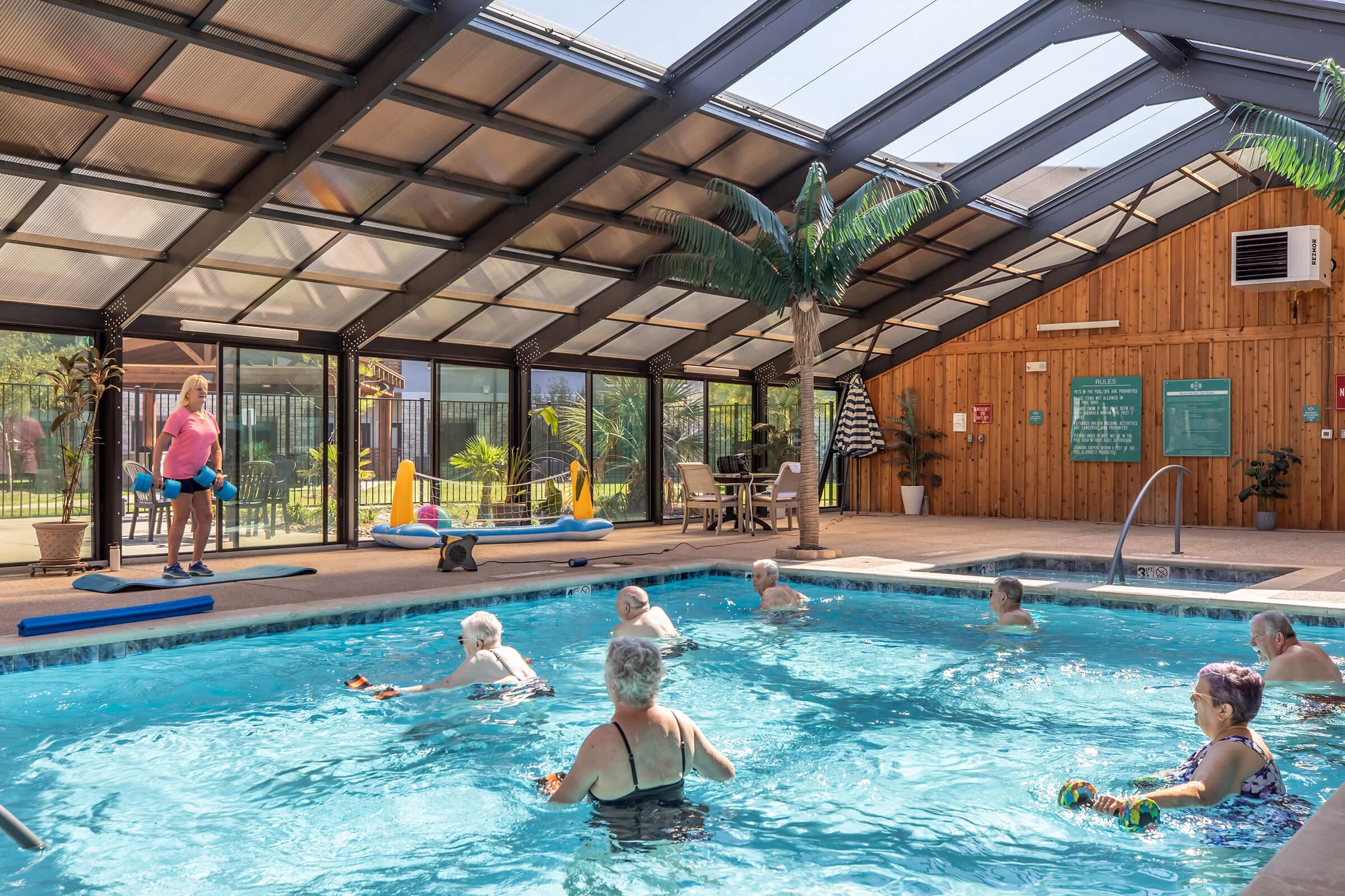 A group of older adults participates in a water aerobics class inside a well-lit indoor pool area. They use water weights while being guided by an instructor. The pool is surrounded by tropical plants, and there is a seating area with tables and lounge chairs.