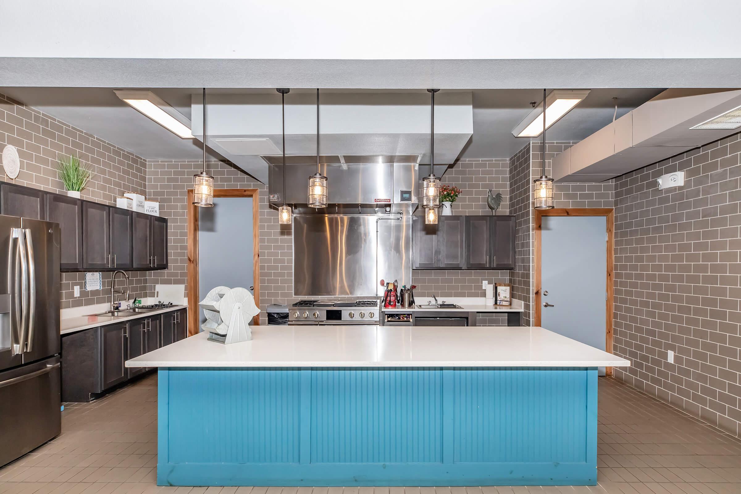 Modern kitchen featuring a large central island with a blue accent, stainless steel appliances, and dark cabinetry. The walls are adorned with gray tiles, and there are pendant lights hanging above the island. The space is well-lit and designed for both functionality and aesthetics.