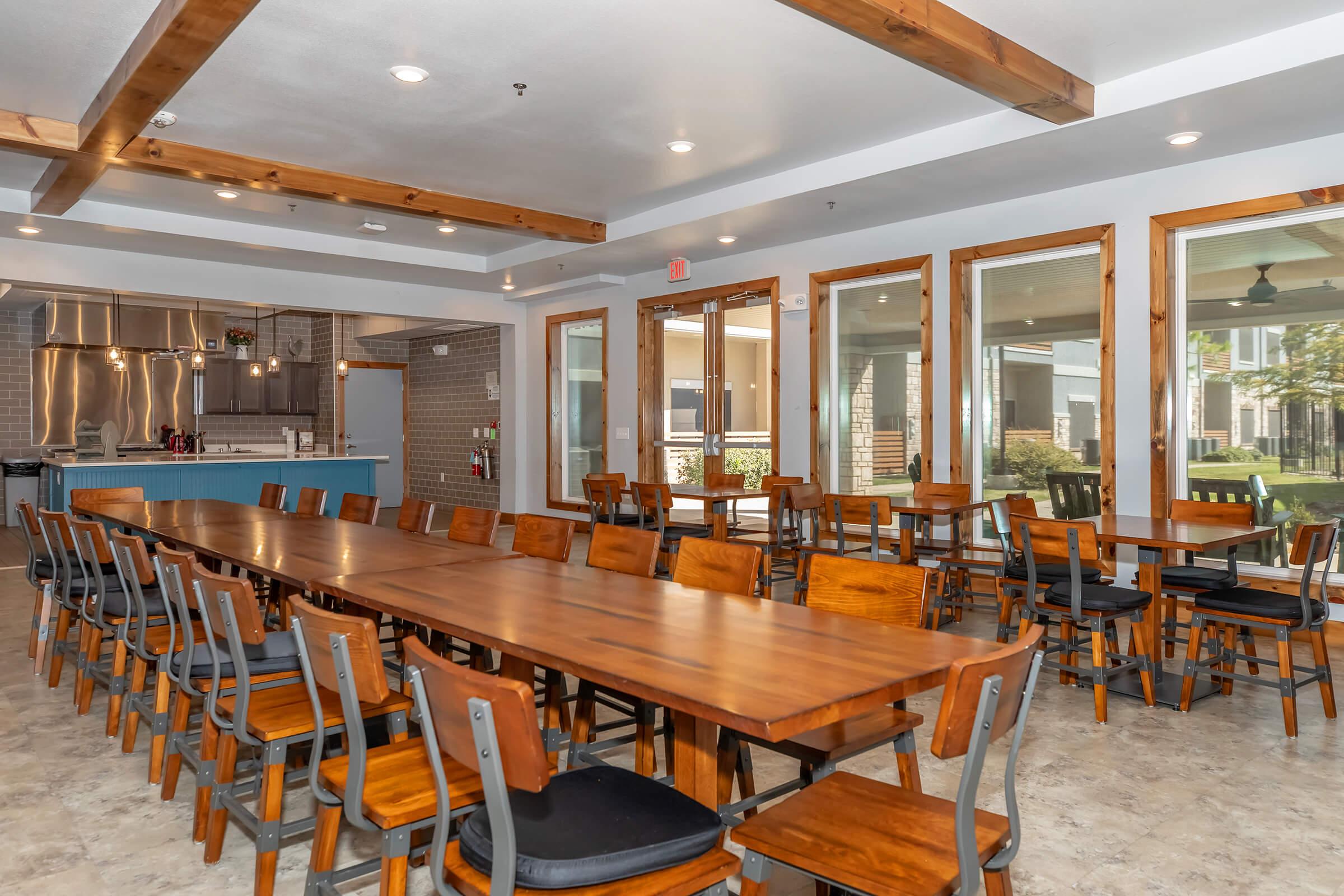 A spacious dining area featuring a long wooden table surrounded by chairs, with large windows allowing natural light to fill the room. The open layout includes a kitchen in the background and offers a view of an outdoor area. Elegant wood beams add a modern touch to the decor.