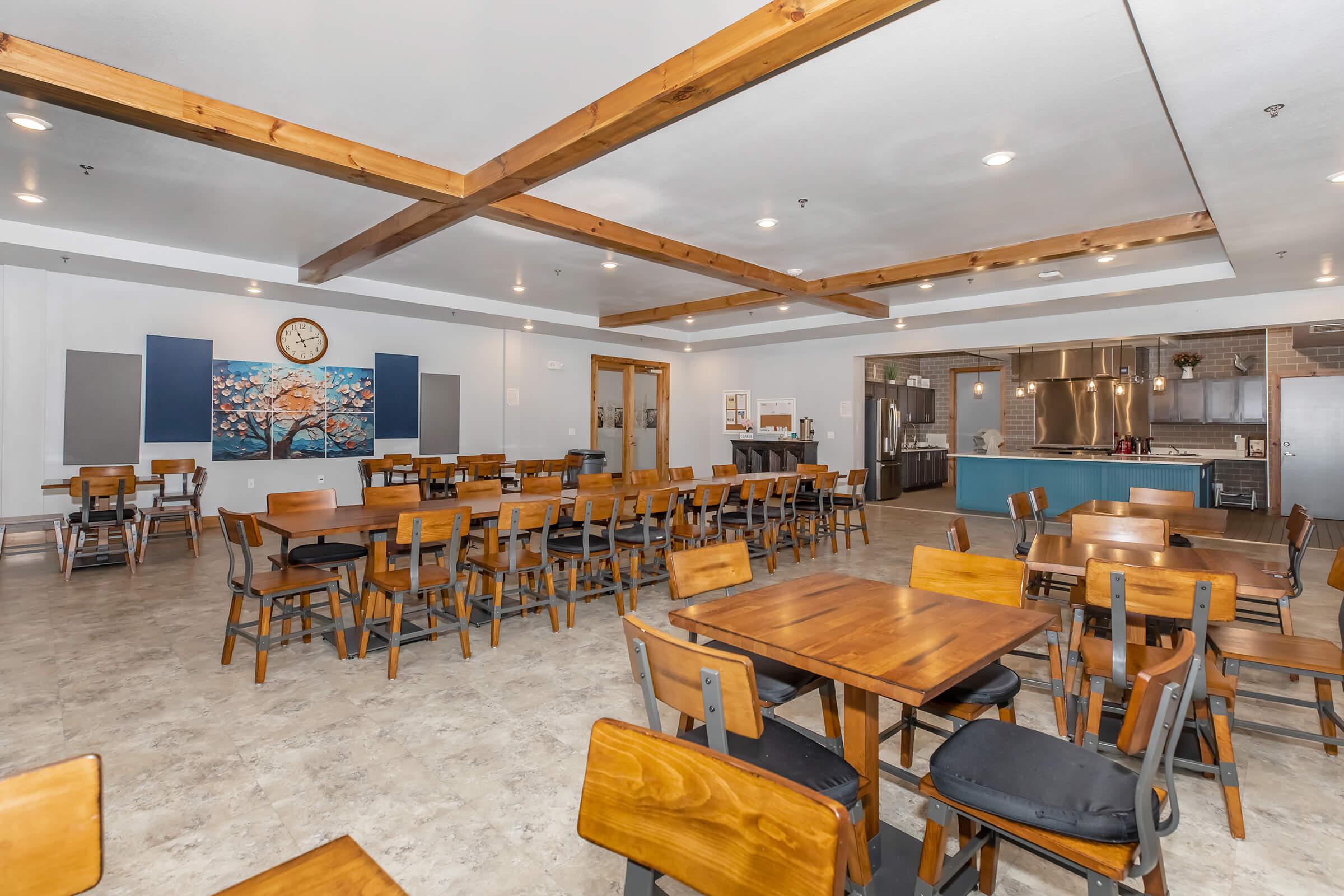 A spacious dining area with wooden tables and chairs arranged neatly. There’s a large kitchen area visible in the background, equipped with modern appliances. A wall-mounted clock is present above a colorful decorative painting. The ceiling features exposed wooden beams, adding to the warm atmosphere.