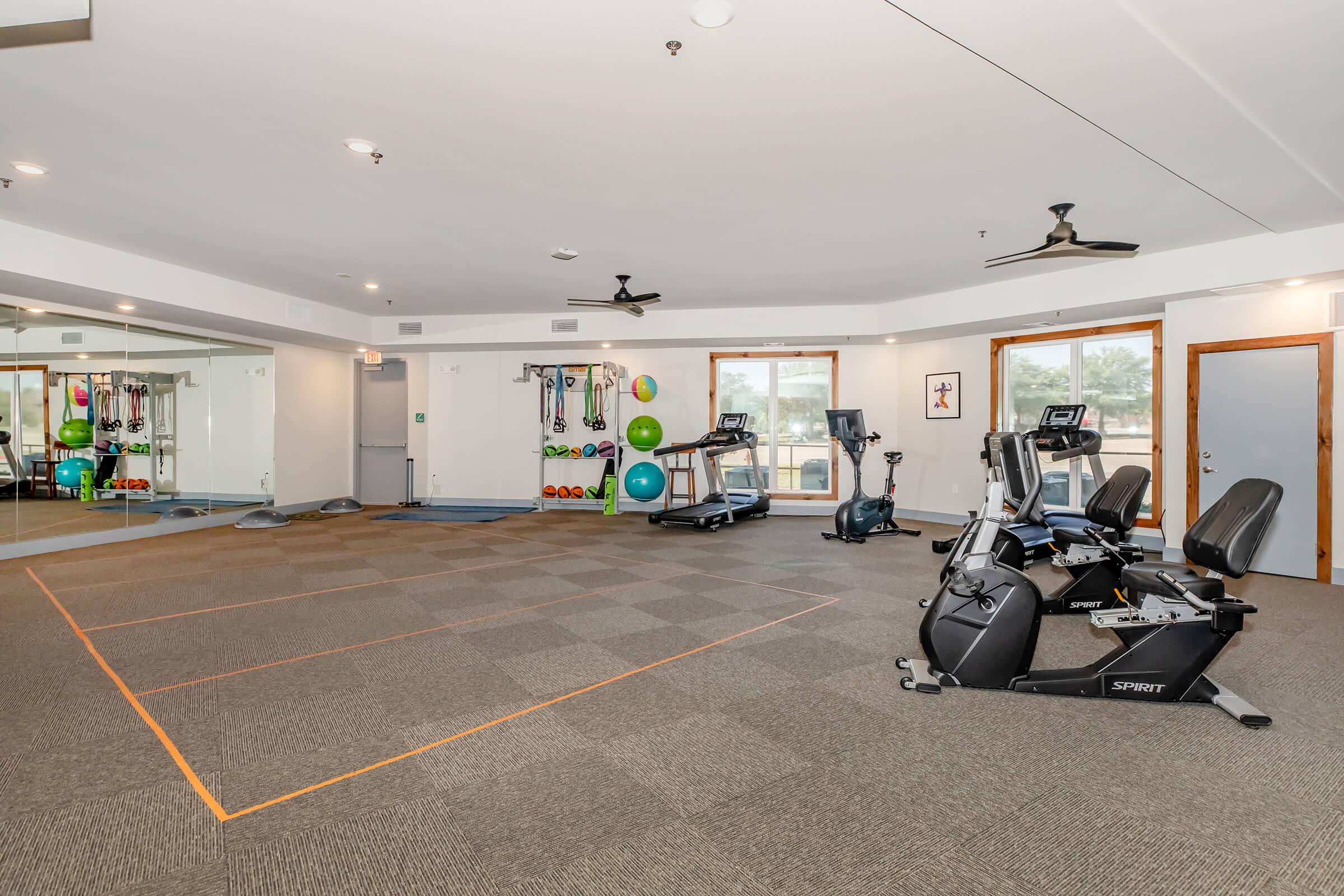 A spacious fitness room featuring various exercise equipment, including stationary bikes and elliptical machines. The area includes colorful exercise balls and resistance bands. Large mirrors on one wall and bright lighting enhance the open atmosphere, perfect for workouts or fitness classes.