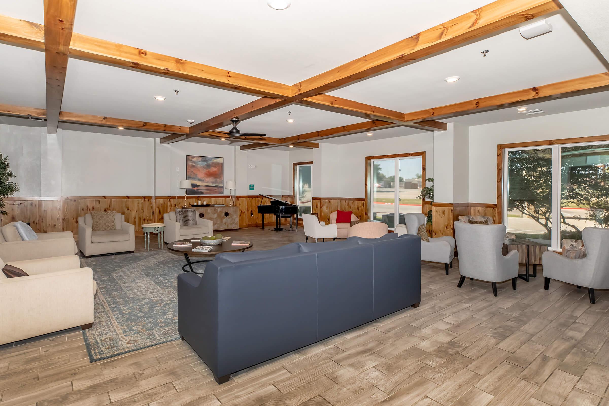 A spacious, modern lounge area featuring a mix of comfortable seating, including sofas and armchairs, with wooden beams on the ceiling. A coffee table is in the center, and large windows allow natural light to fill the room. There is a piano in the background, enhancing the inviting atmosphere.