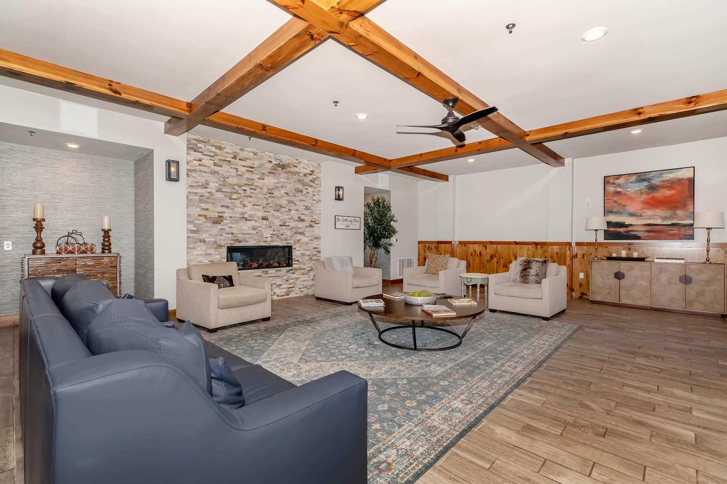A modern living room featuring beige armchairs and a dark blue sofa arranged around a circular coffee table. A stone accent wall with a fireplace is visible, along with wooden beams overhead. A vibrant abstract painting hangs on the wall, and a potted plant adds greenery to the space. Natural light streams in, creating a warm atmosphere.