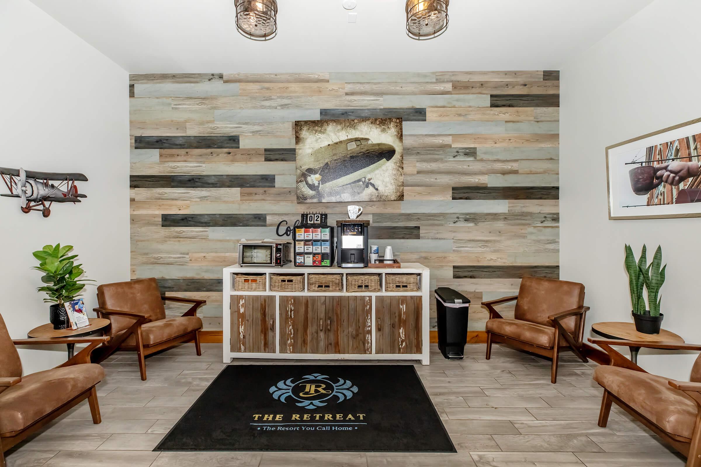 A welcoming lobby area featuring a wood-paneled accent wall, a large artwork of a surfboard, decorative plants, and comfortable brown chairs. There is a wooden console with coffee and snack options, as well as a black mat with "The Retreat" logo on the floor.
