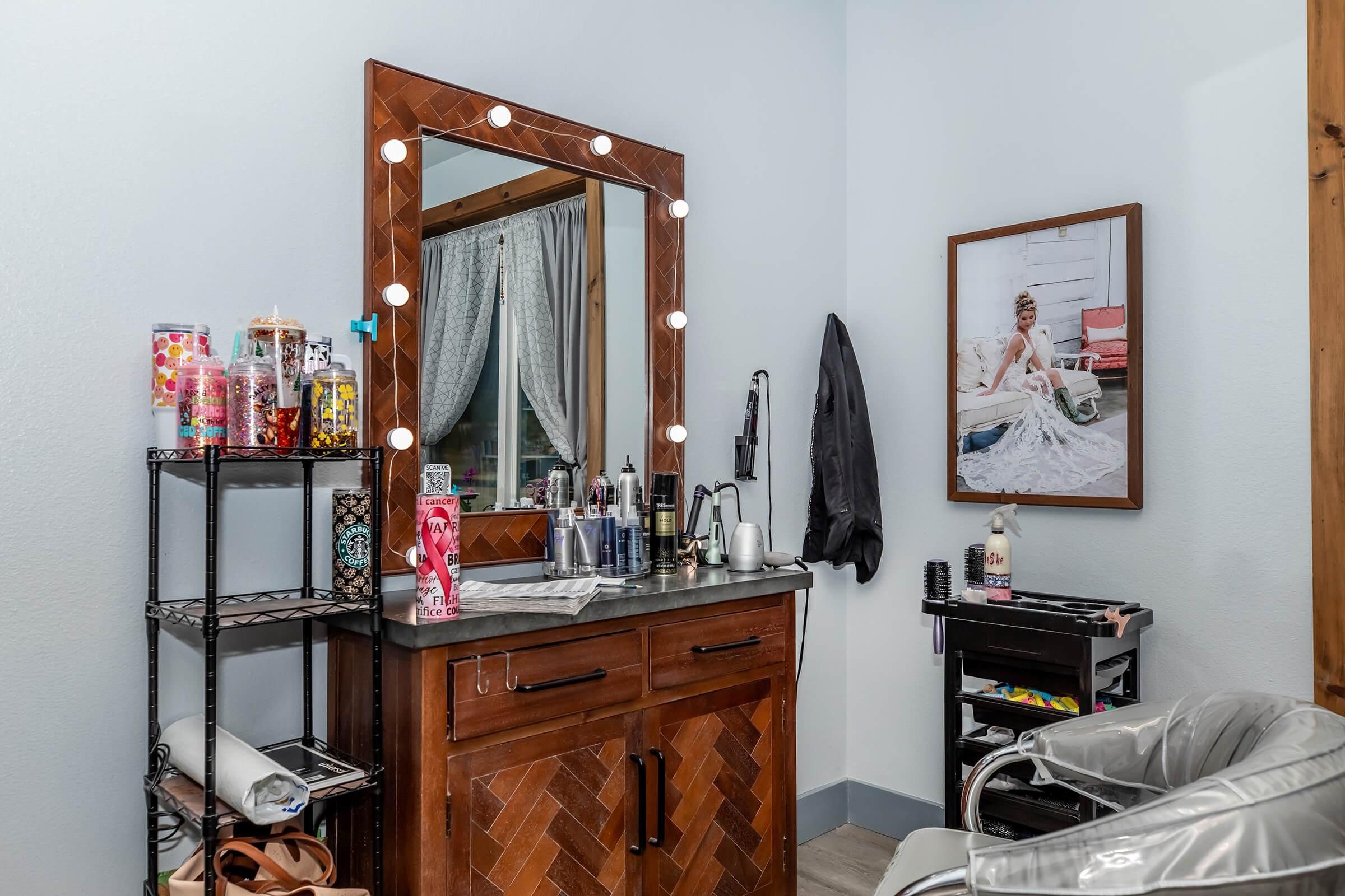 A well-lit salon interior featuring a wooden vanity with a mirror surrounded by light bulbs. There's a shelf with colorful beauty products, and a chair for clients. A framed portrait of a woman in a gown decorates the wall, creating a stylish and inviting atmosphere for hair and beauty services.