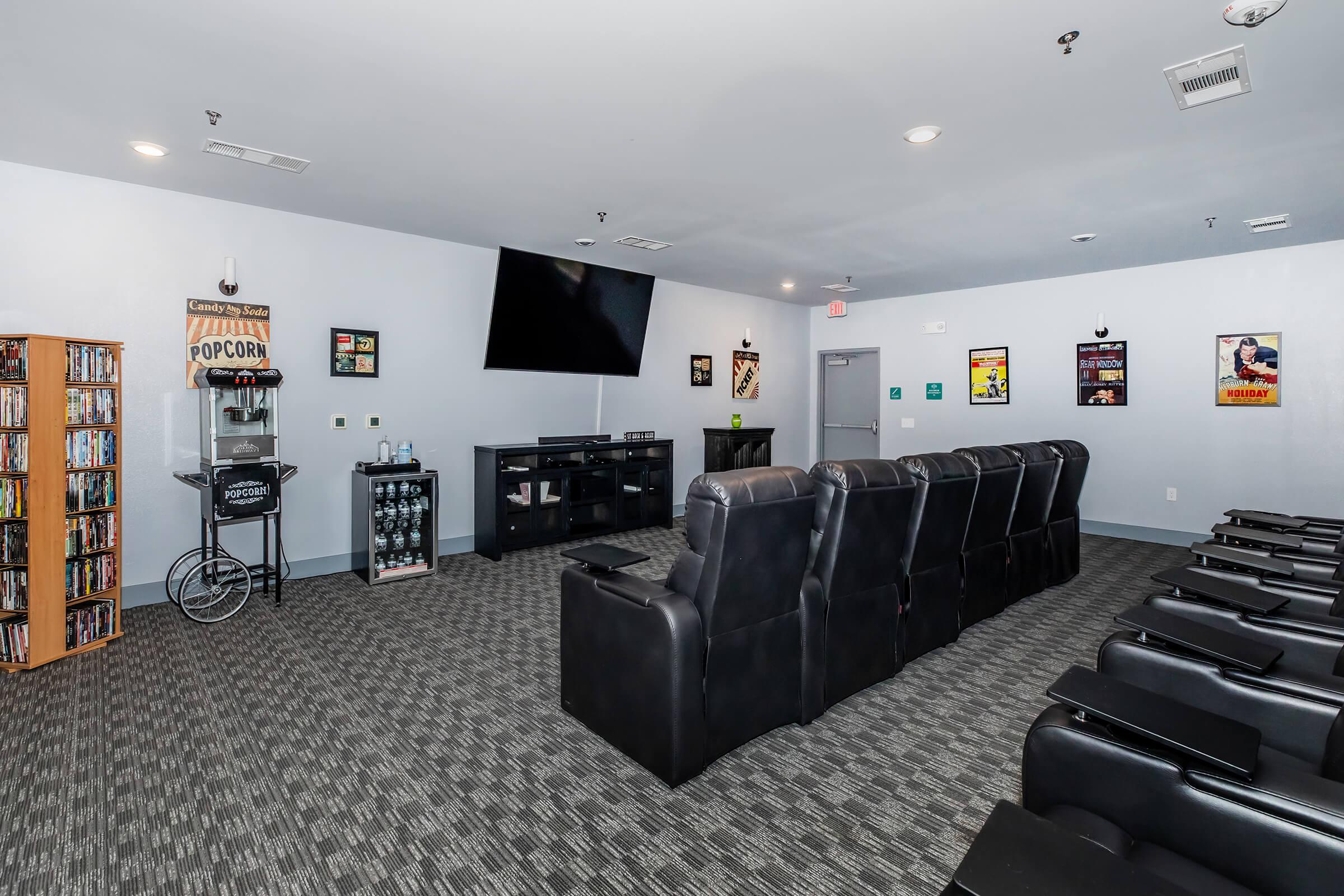 A cozy home theater setup featuring black reclining chairs arranged in front of a large flat-screen TV. To the side, there are shelves filled with DVDs and a popcorn machine, complemented by nostalgic movie posters on the walls, creating an inviting movie-watching atmosphere.