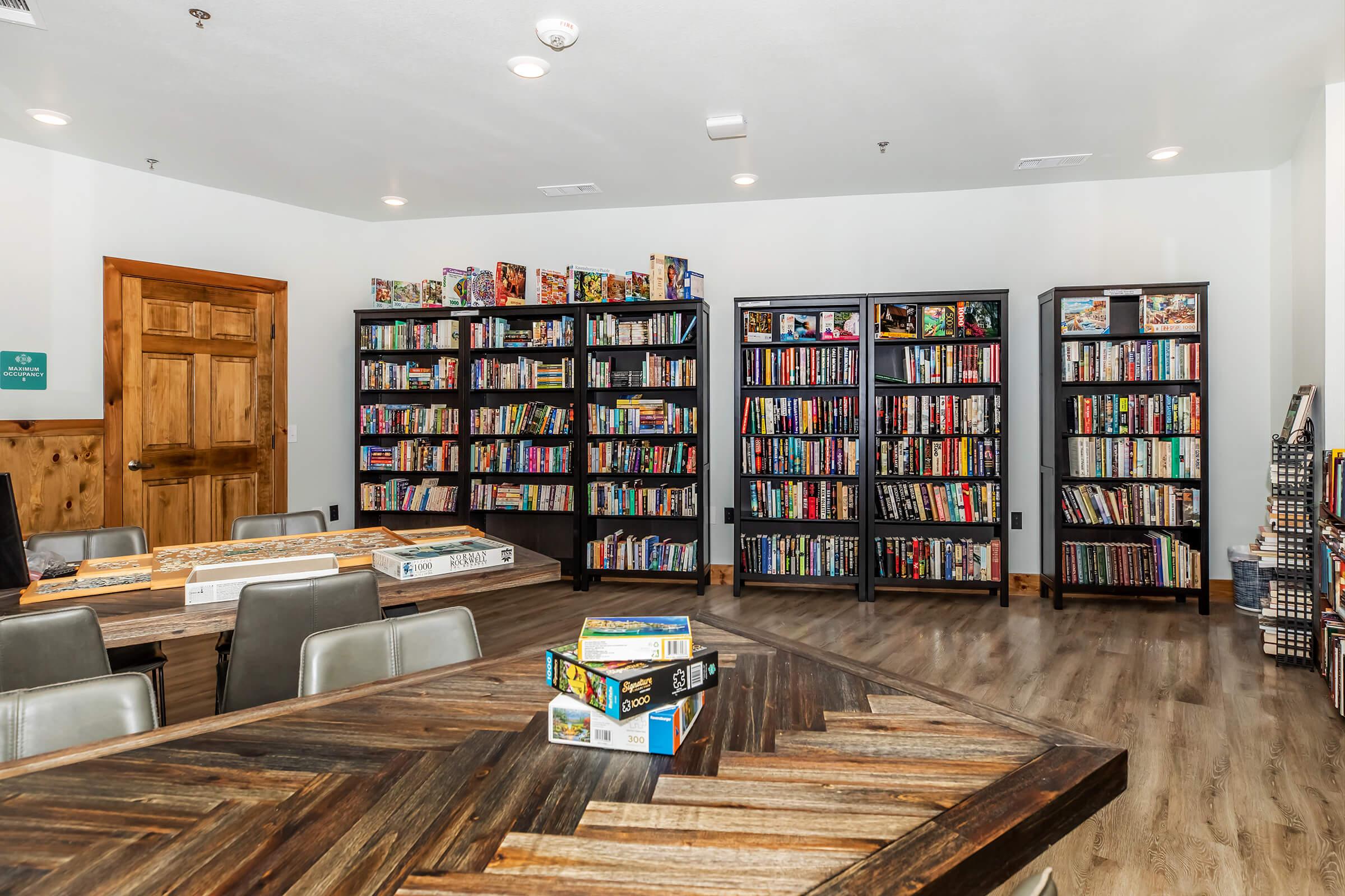 A cozy library or game room featuring several tall bookshelves filled with books and board games. In the foreground, a wooden table with a unique design is surrounded by chairs. The space is well-lit and has a welcoming atmosphere, ideal for reading or playing games.